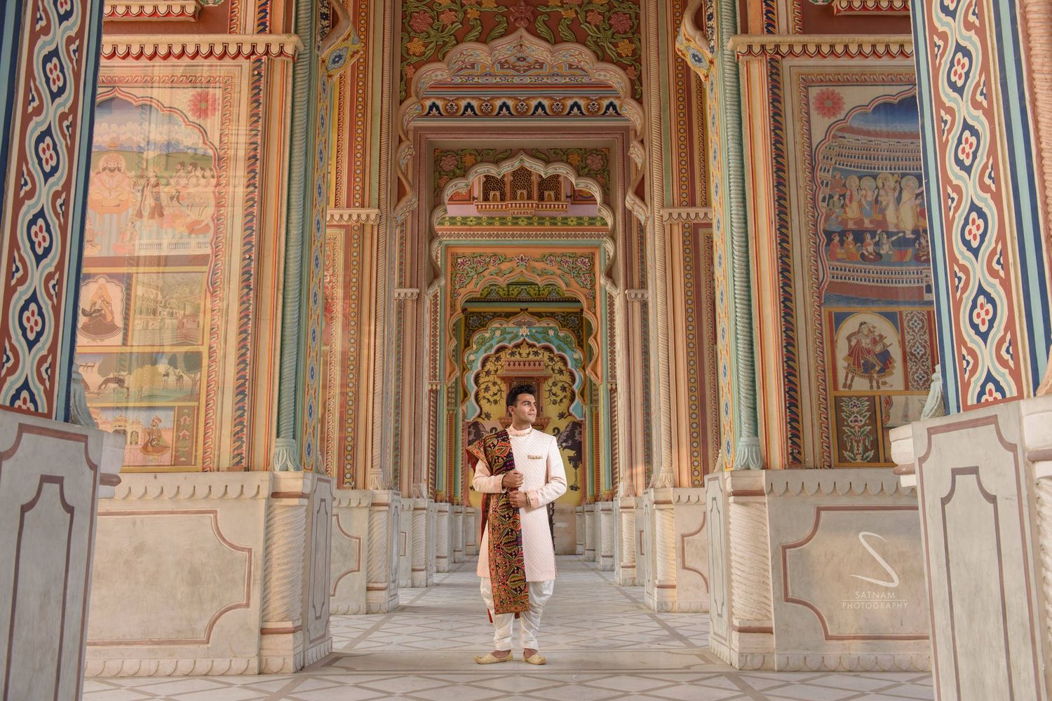 Man in traditional Indian white kurta standing in colorful ornate painted temple hallway.