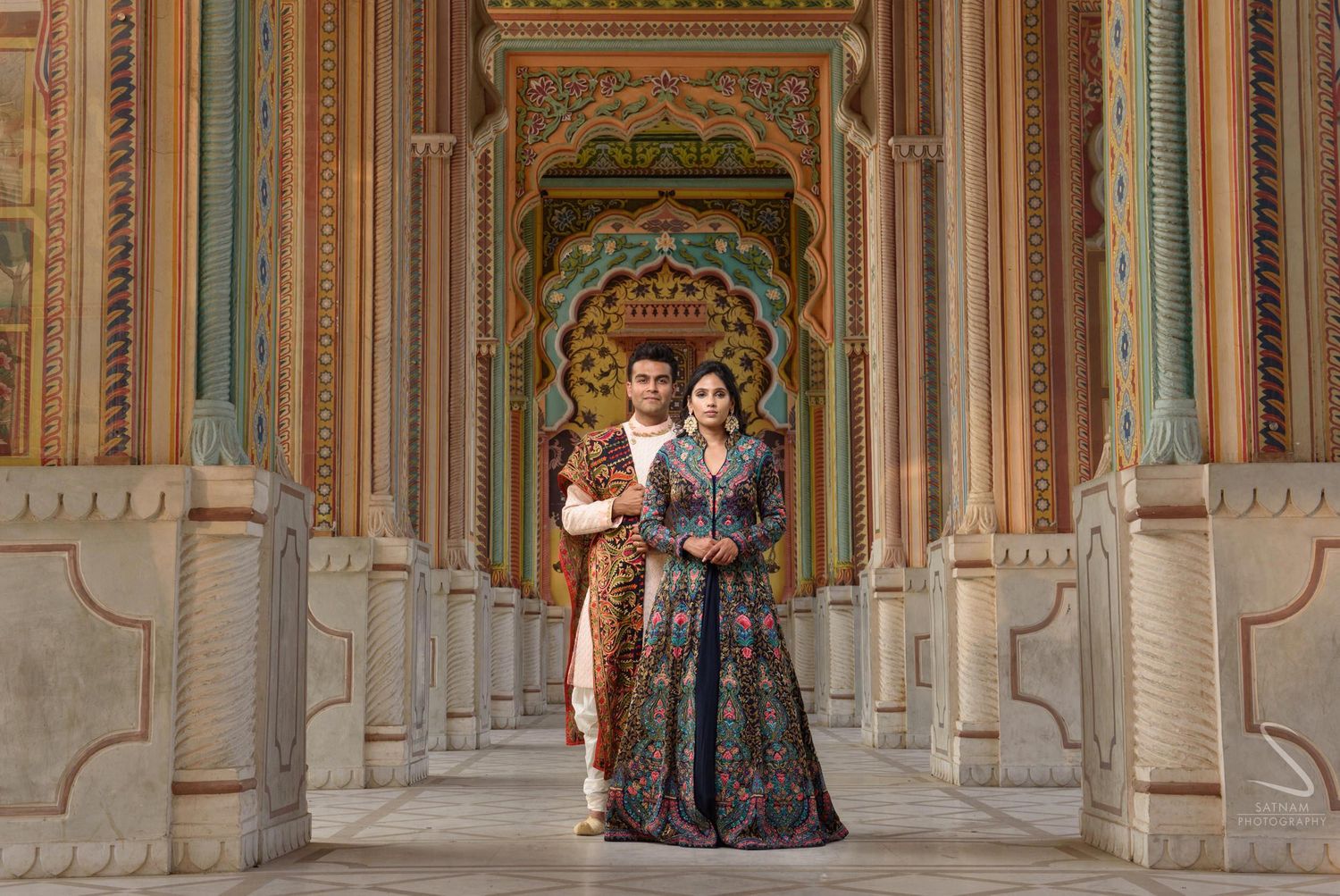 Couple in traditional Indian attire posing in ornate colorful palace corridor in Jaipur.
