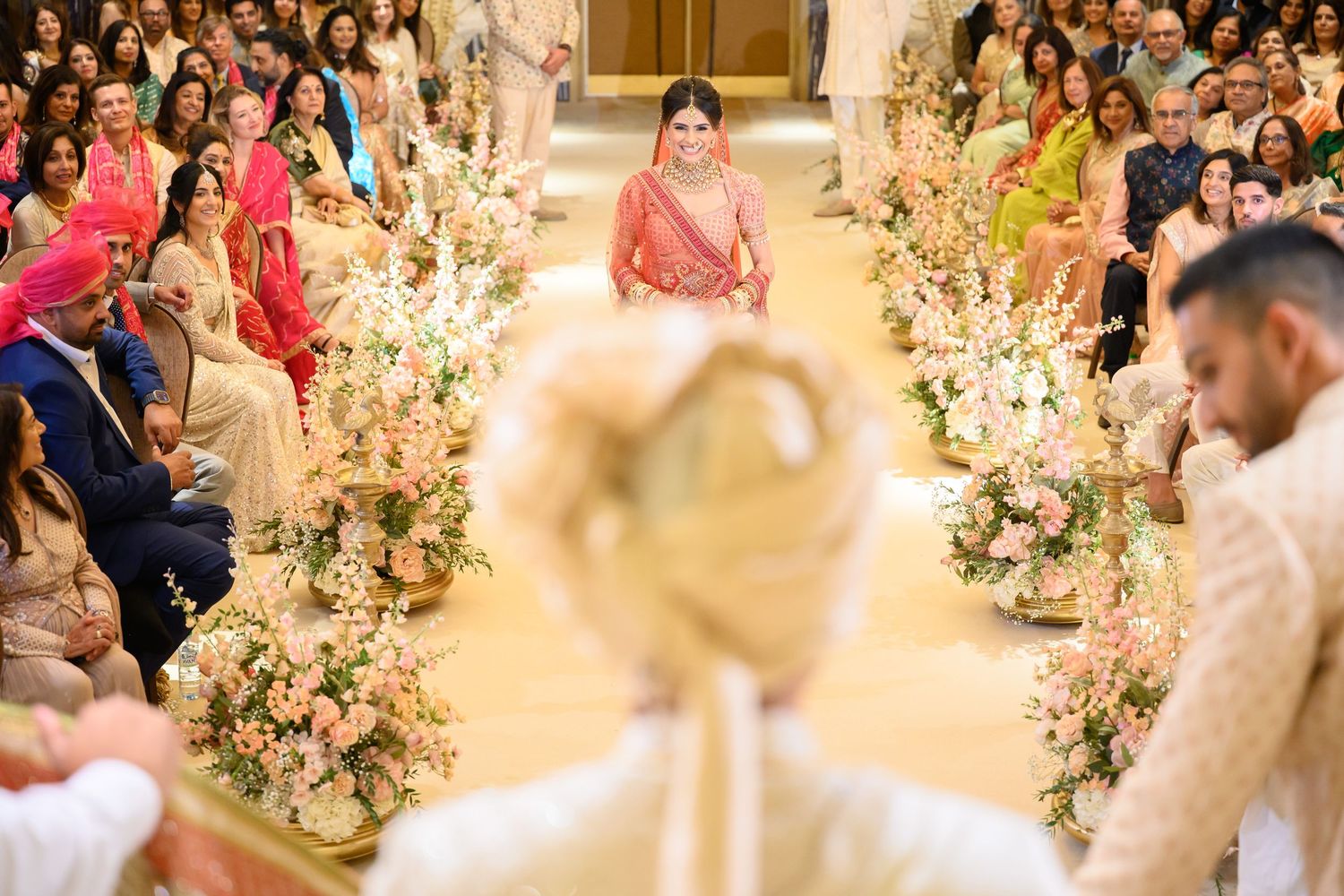 Indian bride walking down floral aisle in pink bridal lehenga during Sikh wedding ceremony.