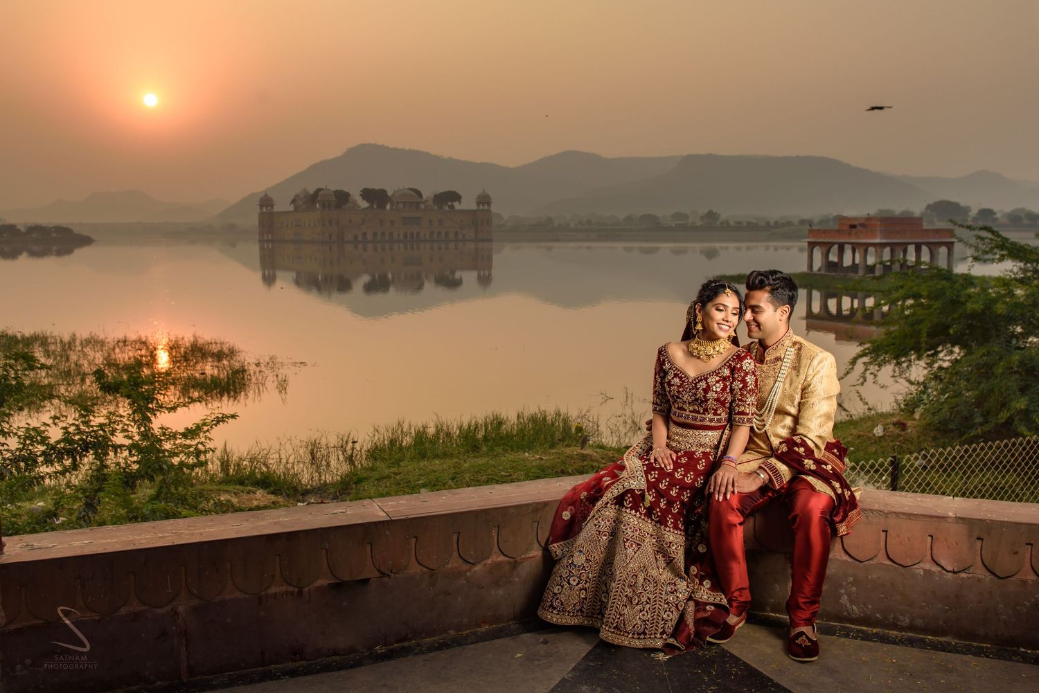 Couple in rich red Indian bridal attire sitting romantically by a lake with Jal Mahal palace at sunset.