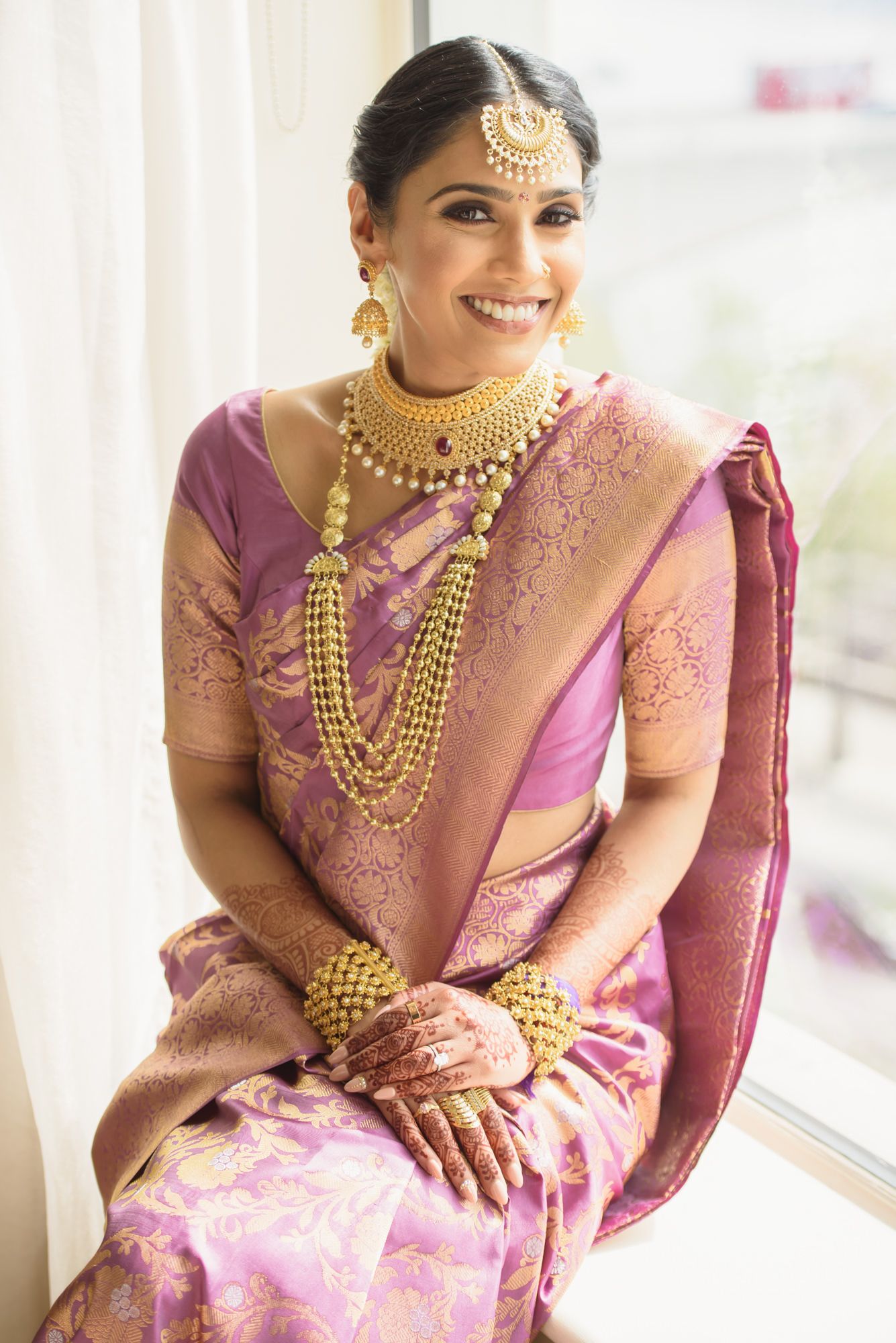 Beautiful Indian bride in purple silk saree with gold jewelry and mehndi smiles on her wedding day.