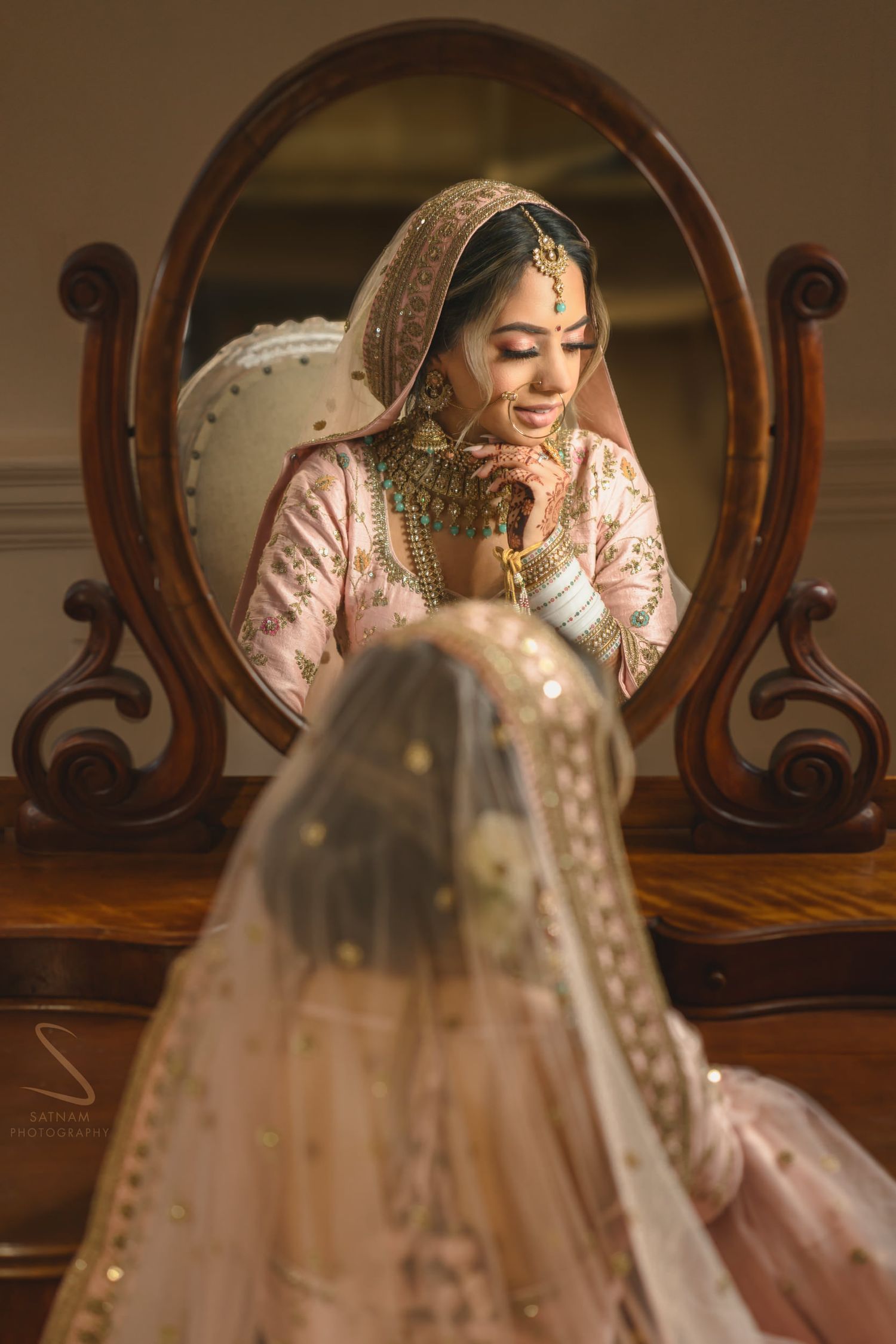 Sikh bride in blush pink lehenga smiling at reflection in ornate vintage mirror.