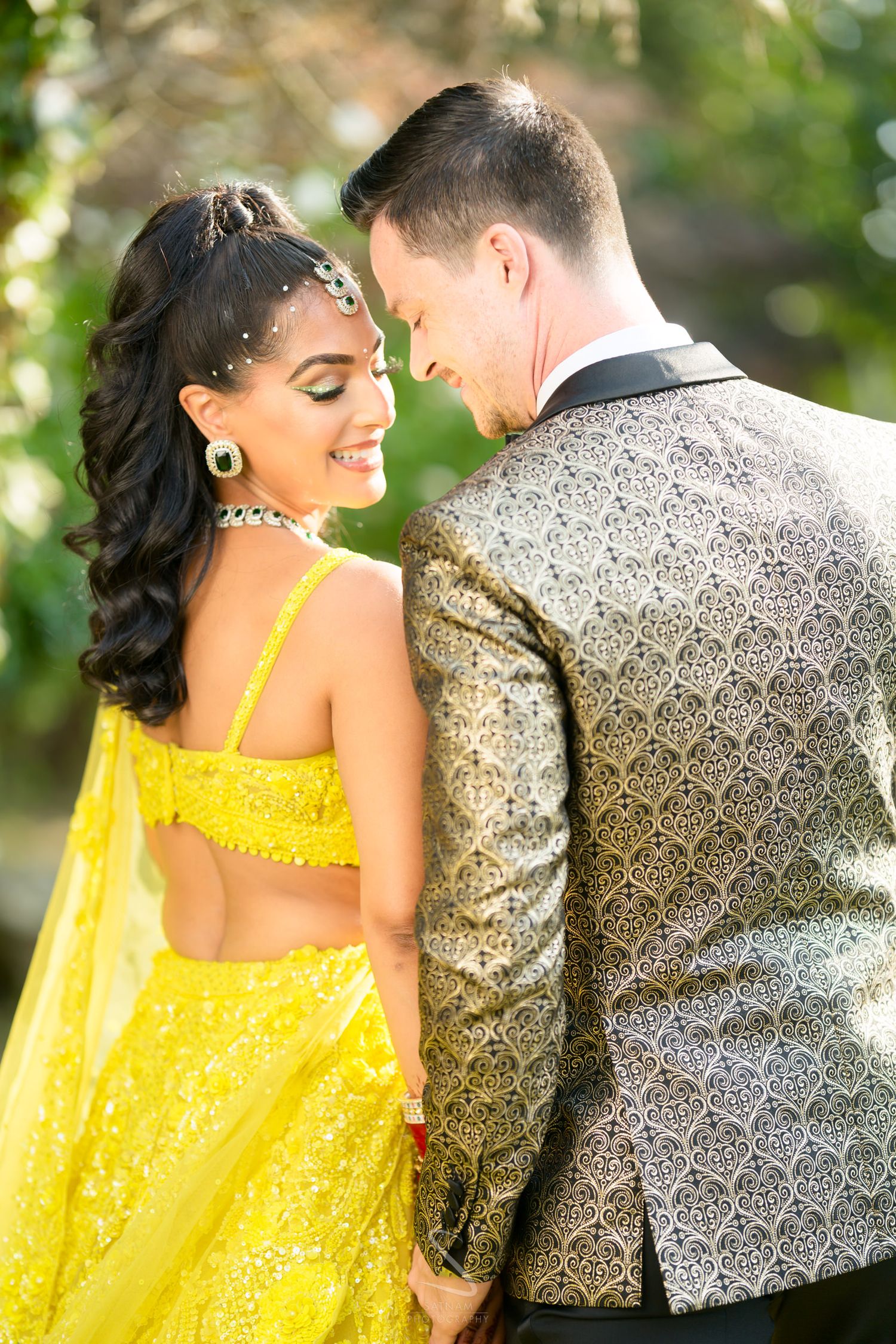 Indian bride in yellow lehenga and groom in gold brocade jacket sharing romantic moment outdoors.