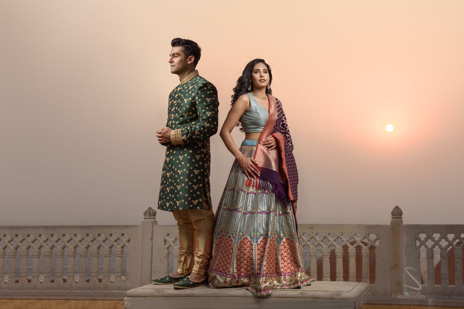 Indian couple in traditional bridal wear posing at sunset, woman in colorful lehenga and man in green sherwani.
