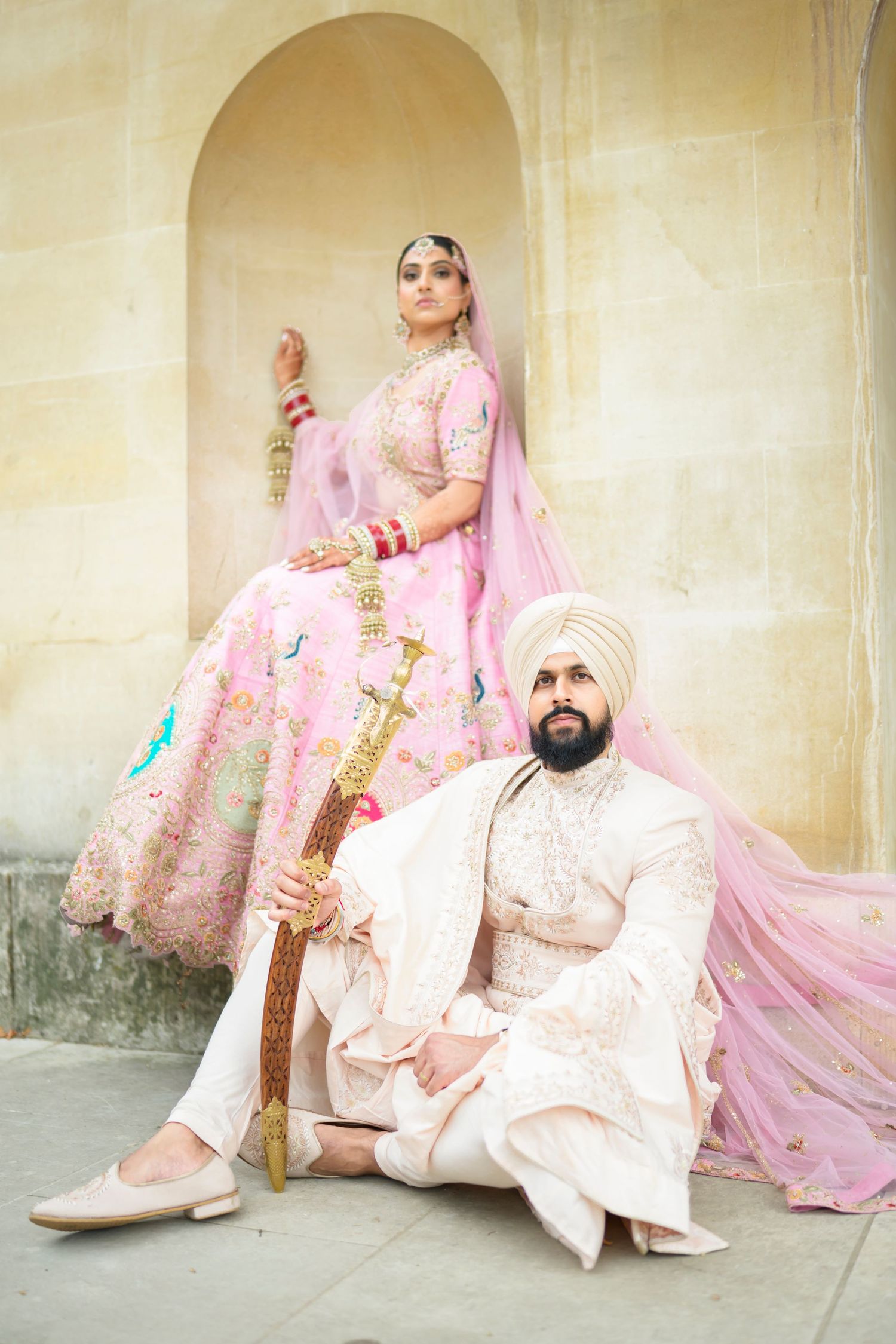 Sikh bride in pink lehenga and groom in white sherwani posing elegantly with ceremonial sword at heritage venue.