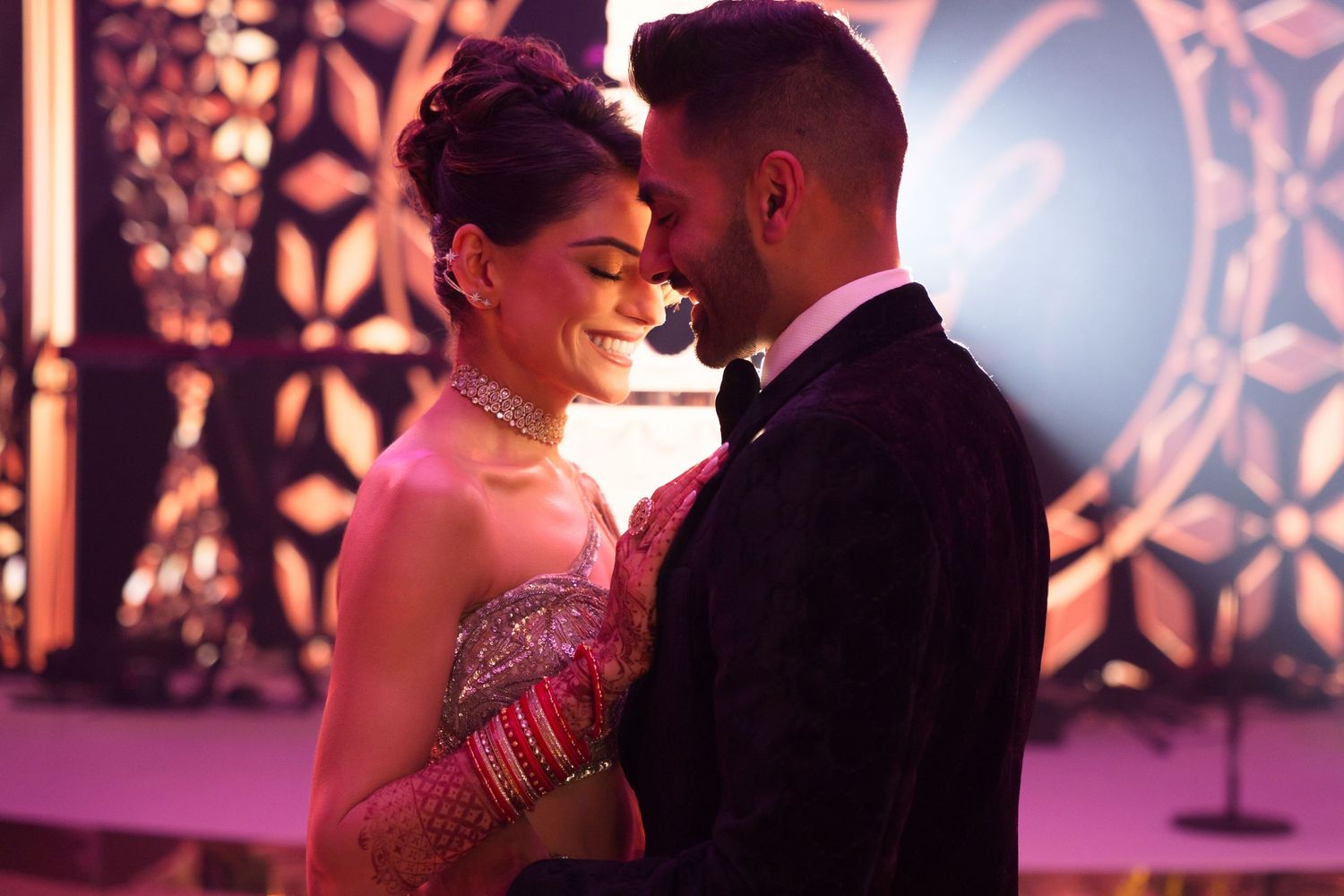 Couple in elegant Indian wedding attire sharing a romantic moment under warm pink lights.
