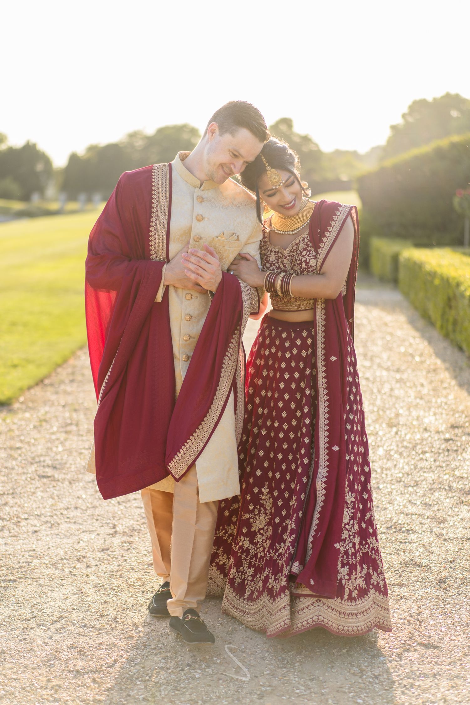 A couple in matching burgundy and gold Indian wedding attire walk together along a sunlit garden pathway.