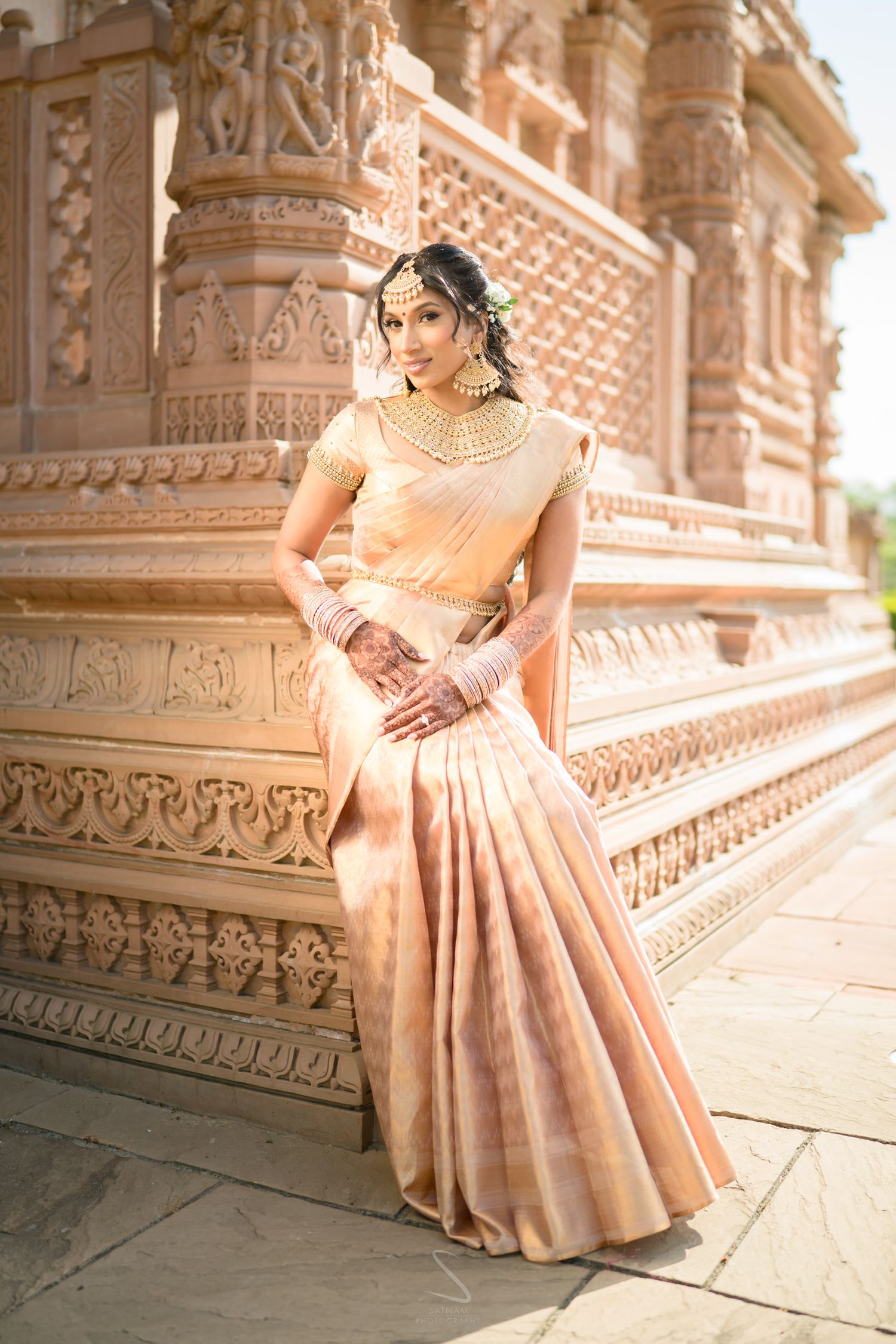 South Asian bride in peach silk saree with gold blouse poses beside ornate temple stone carvings.