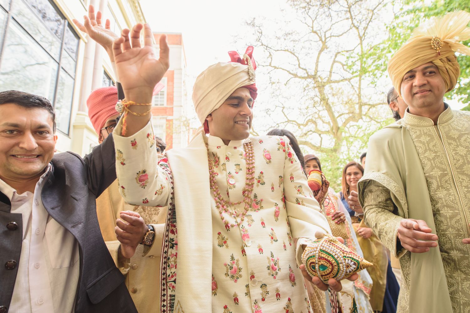 Groom in cream sherwani and gold turban waves during Indian wedding baraat procession outdoors.