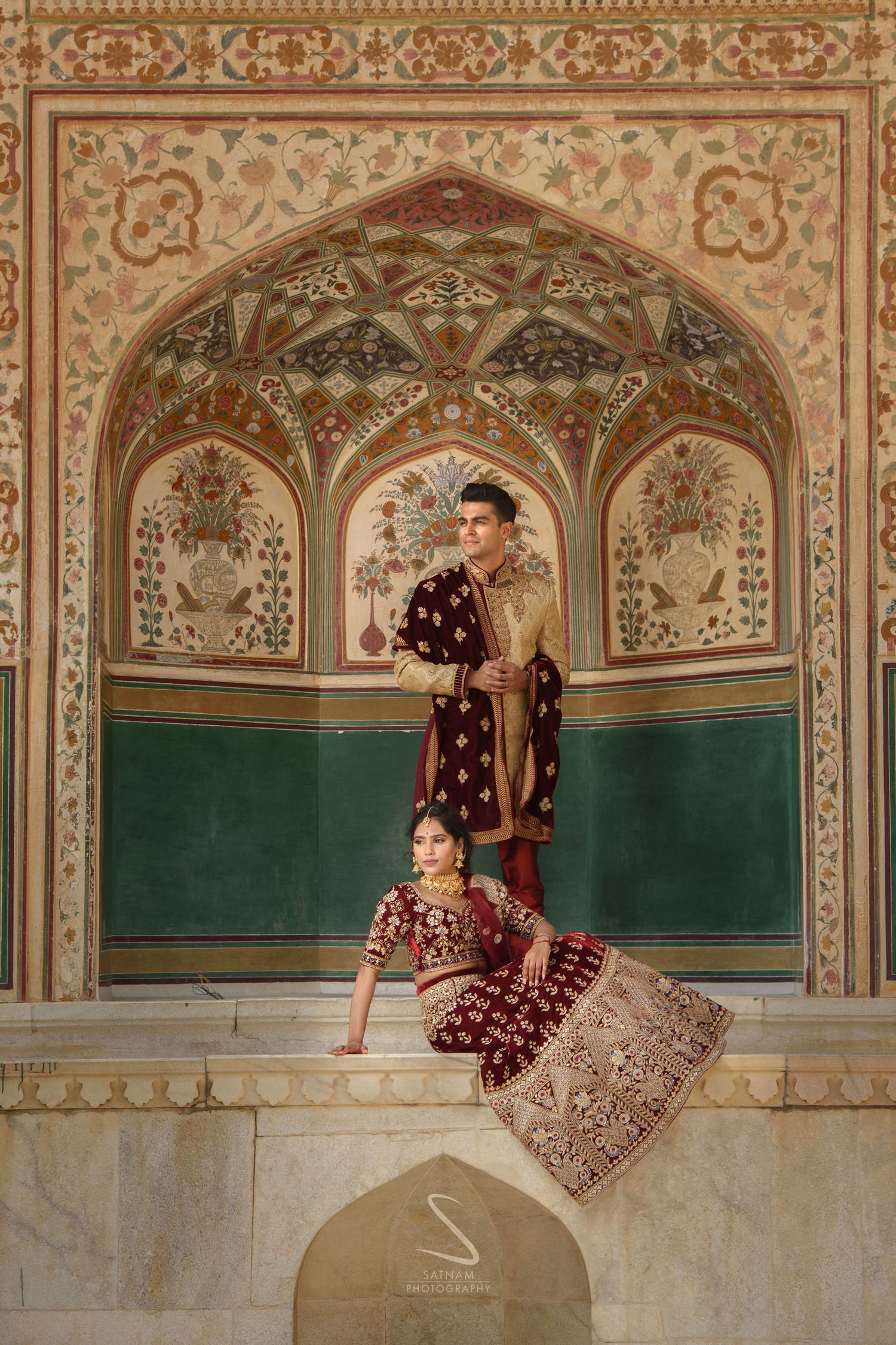 Indian bride in maroon lehenga and groom in sherwani posing in ornate Amber Fort archway, Jaipur.