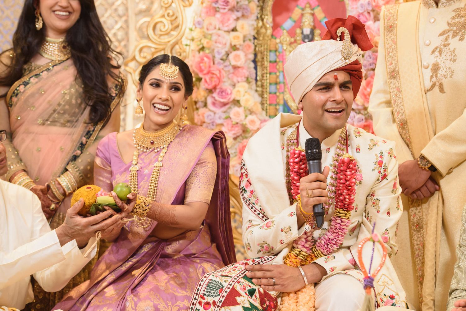 Joyful Indian bride in purple saree and groom in white sherwani laugh during traditional wedding ceremony.