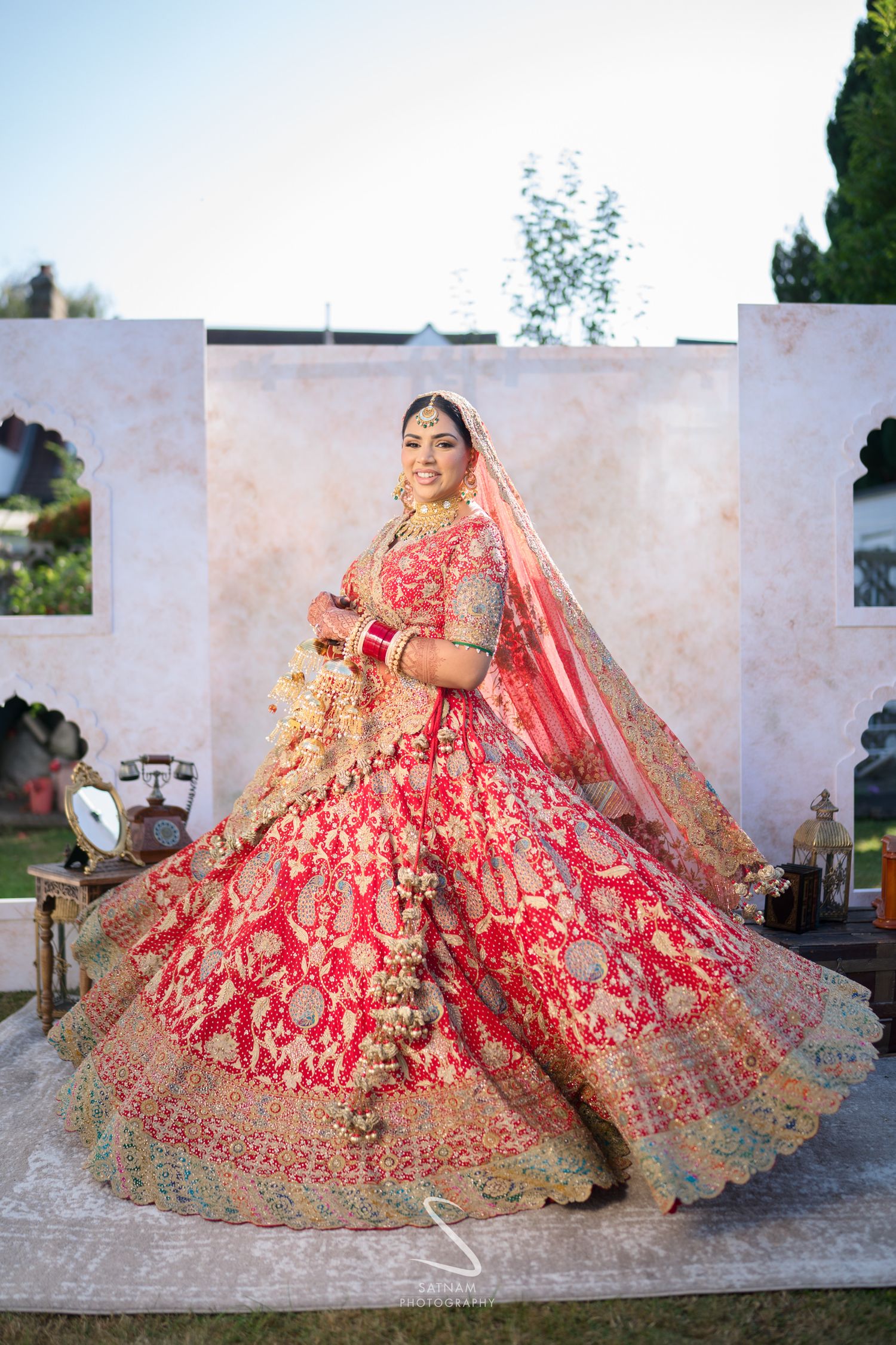 Indian bride twirls in stunning red heavily embroidered bridal lehenga with gold detailing at outdoor venue.