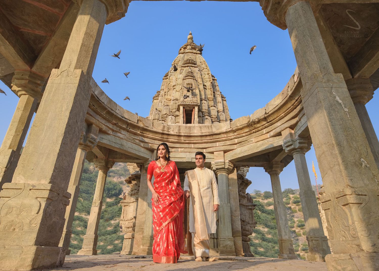 Indian couple in red saree and kurta walking through ancient temple pillars in Rajasthan.