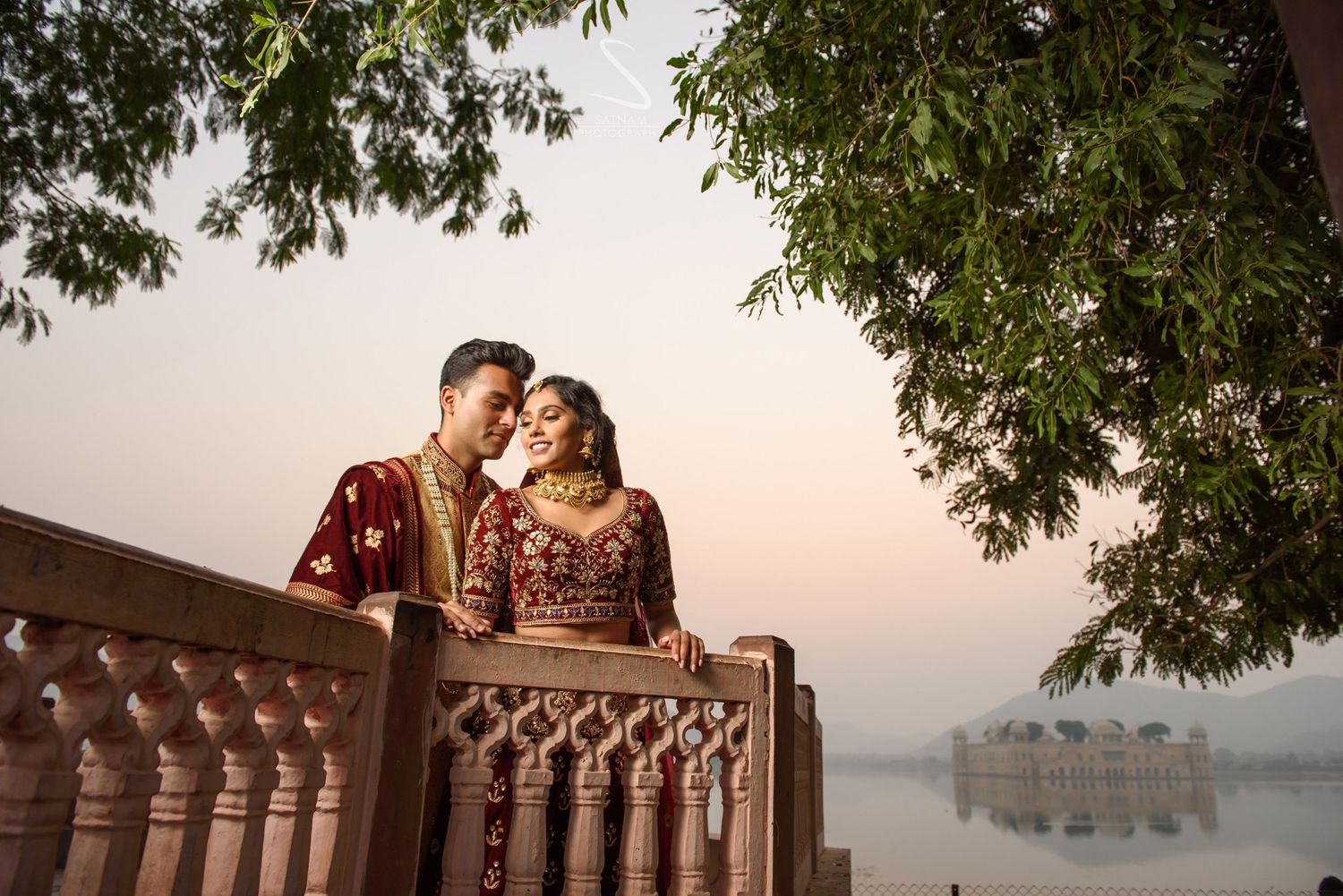 Romantic couple in red Indian bridal wear embracing on a wooden balcony overlooking a misty lake.