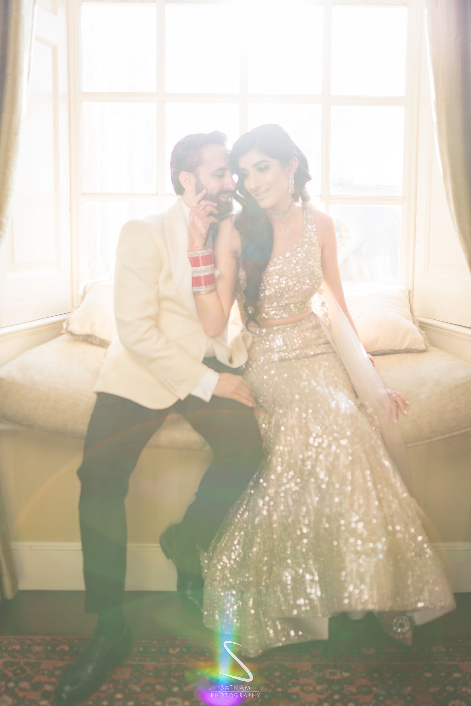 An Indian bride in a gold sequined lehenga and groom in a white suit share an intimate moment by a sunlit window.