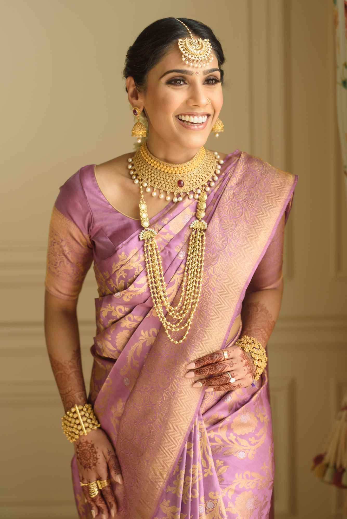 Smiling South Indian bride in purple silk saree with traditional gold bridal jewelry set.