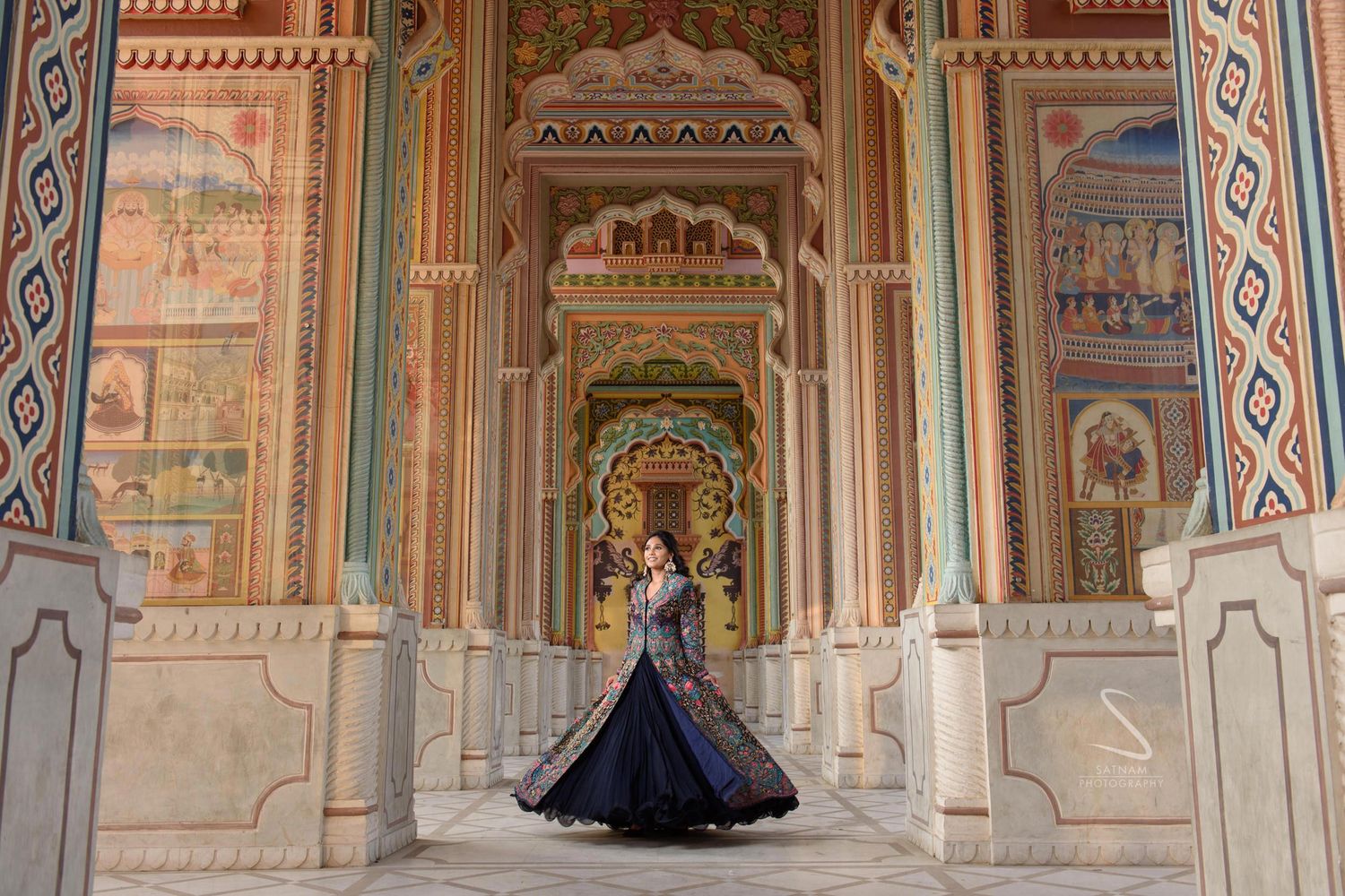 Woman in elegant navy lehenga twirling in vibrant ornately painted Indian temple corridor.