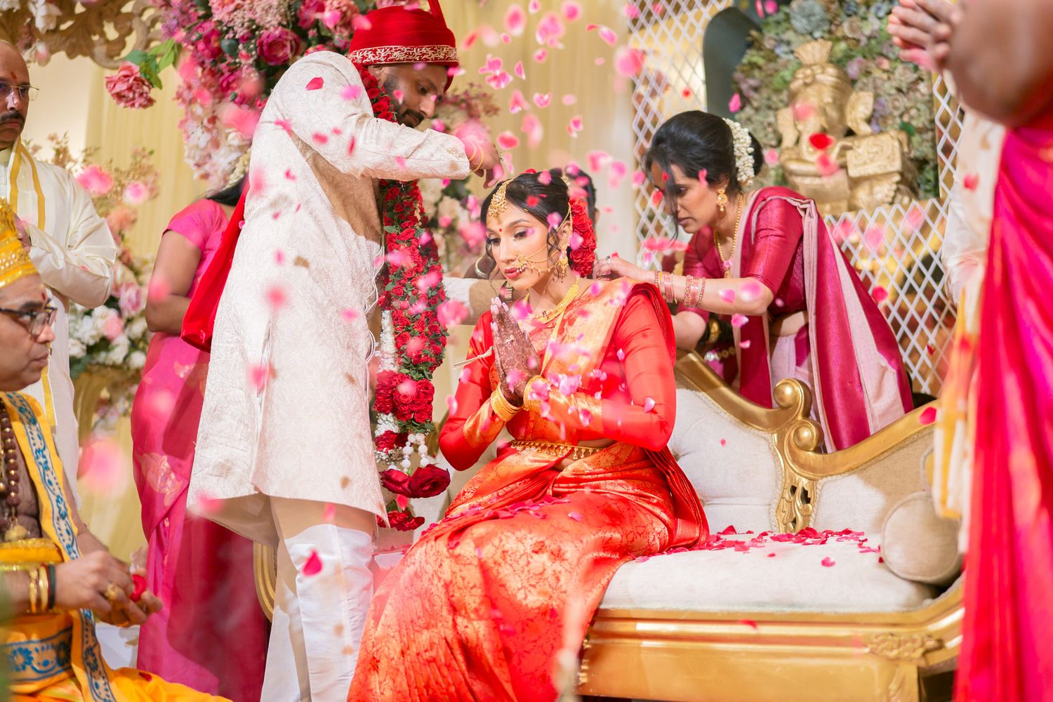 Hindu bride in red saree sits during traditional wedding ceremony with flower petals being showered.