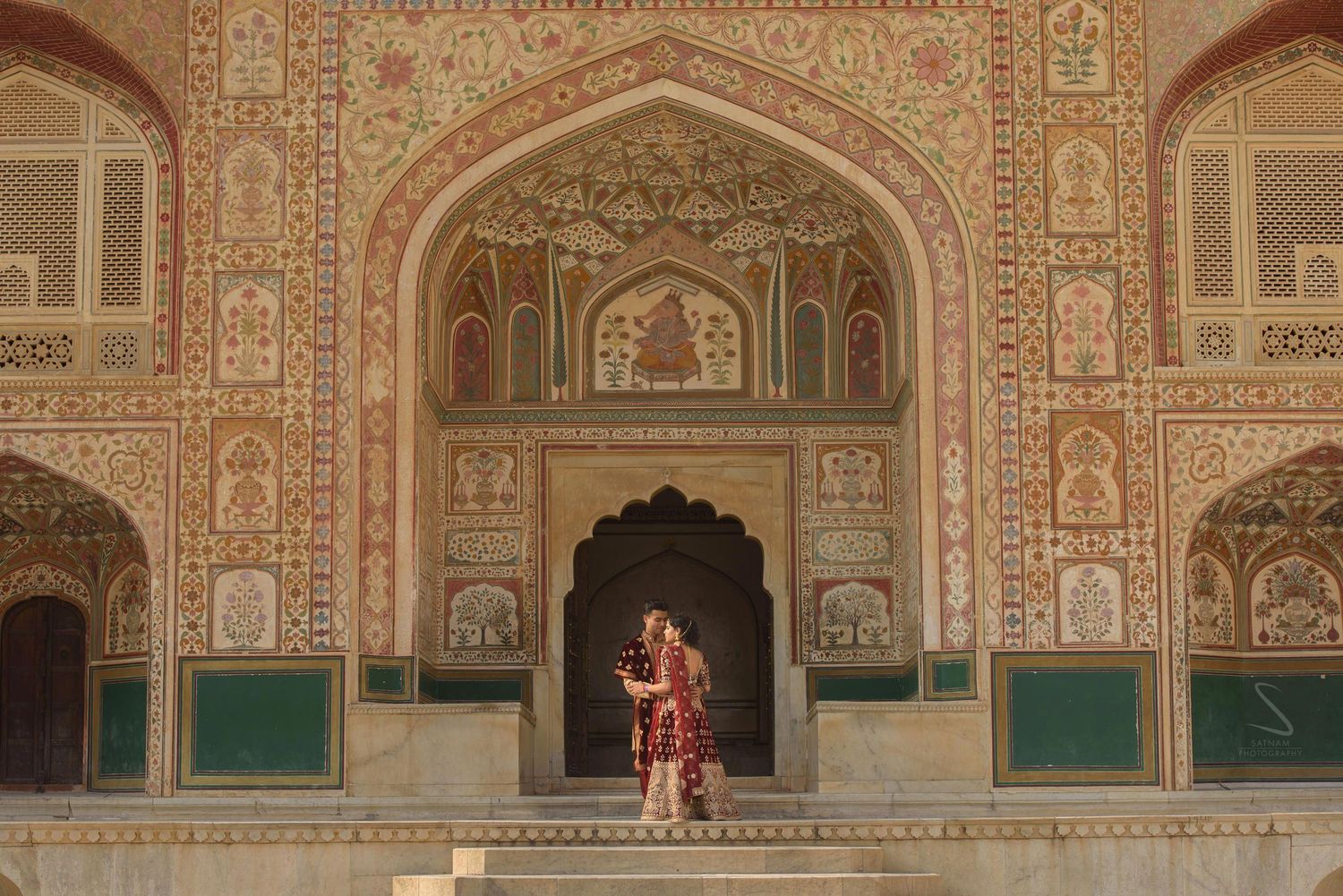 Bride in traditional red lehenga poses under ornately carved and painted arches of Amer Fort.
