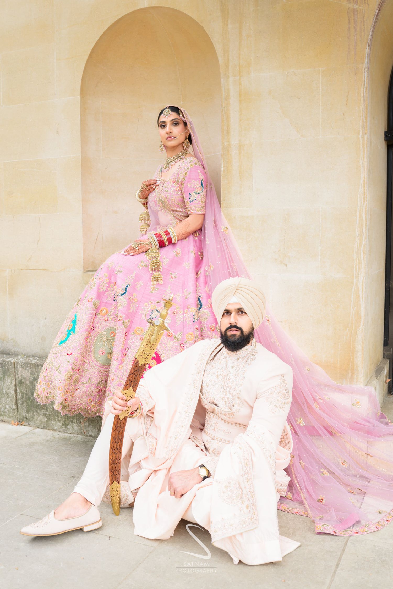 A Sikh bride in a pink lehenga with dupatta stands above her groom in white sherwani against a stone arch backdrop.