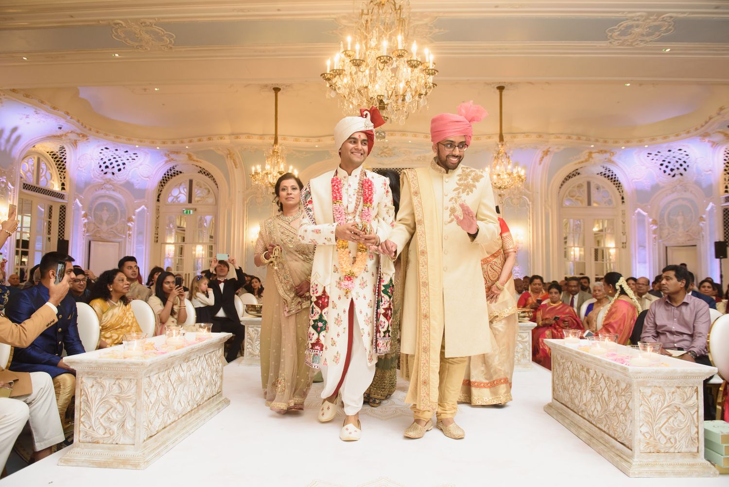 Indian bride and groom walk down aisle in elegant ballroom during traditional Hindu wedding ceremony.