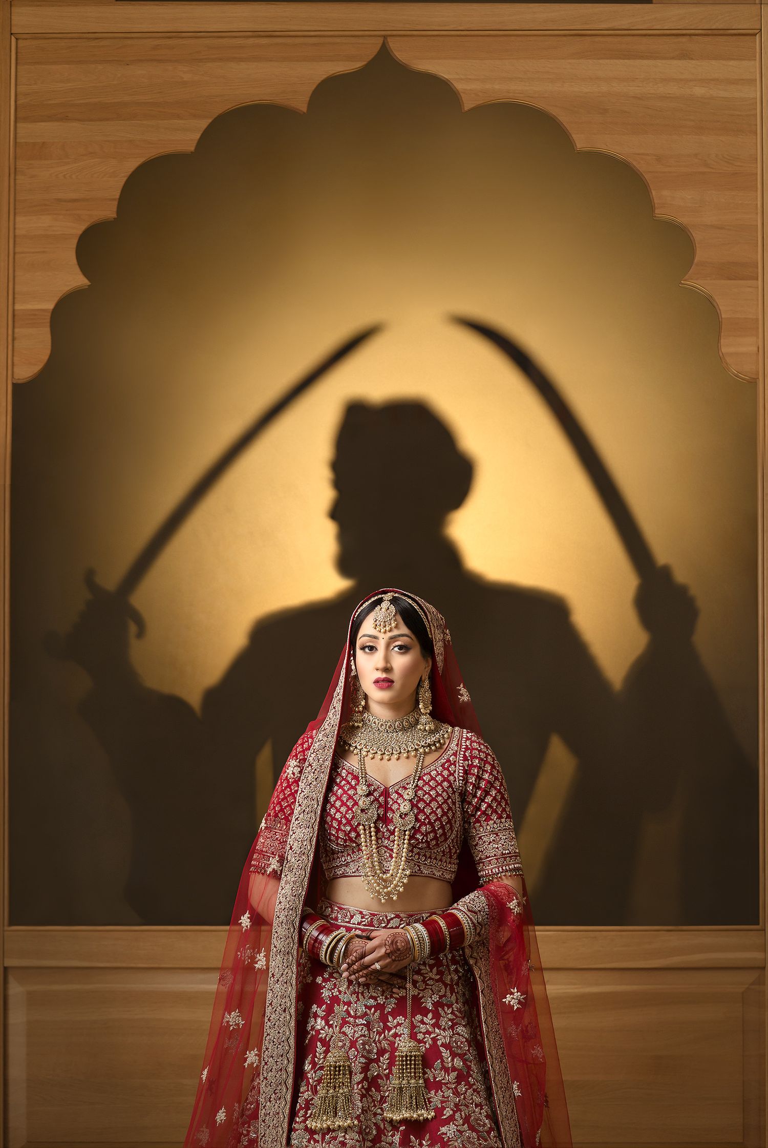 Indian bride in red bridal lehenga standing before dramatic shadow silhouette of sword-wielding warriors.