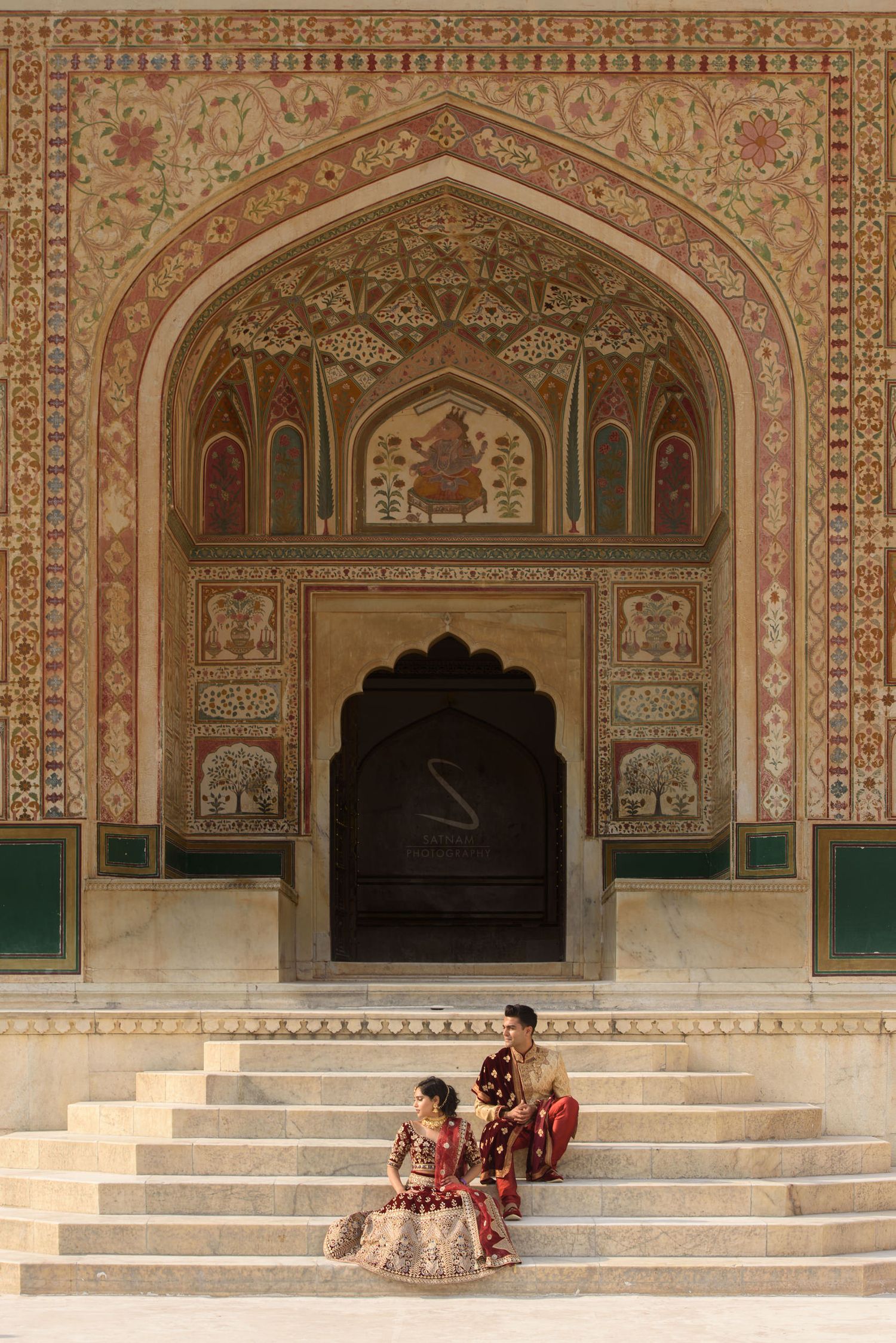 Indian bride and groom in maroon wedding outfits sitting on steps of ornate Mughal archway at Amer Fort.