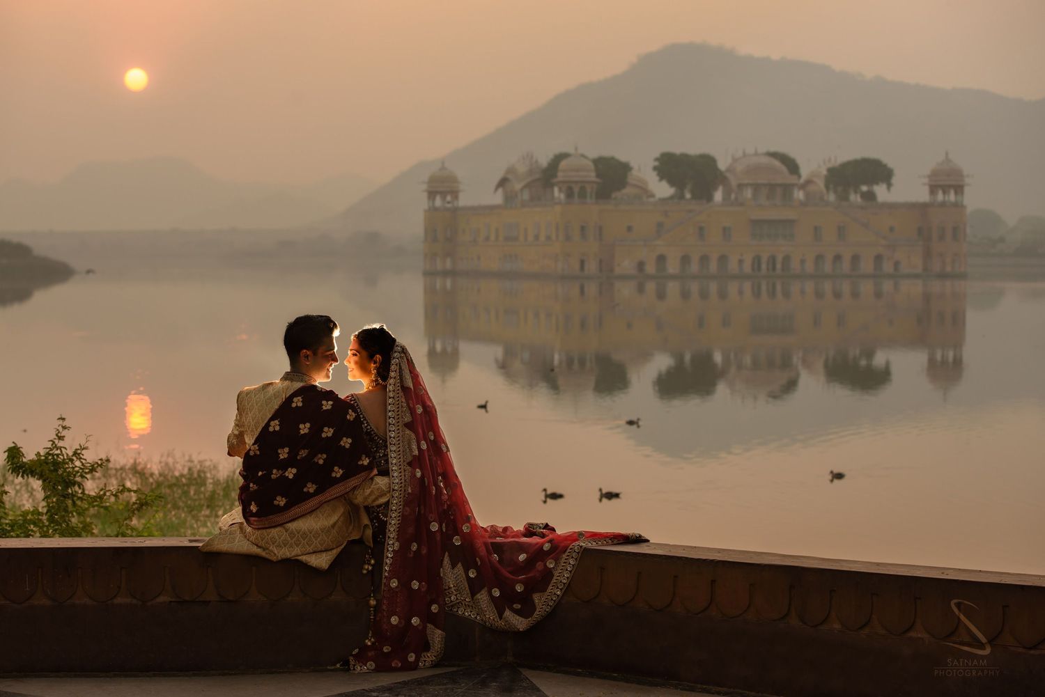 Indian couple in traditional attire sitting by Jal Mahal palace lake at misty sunset in Jaipur.