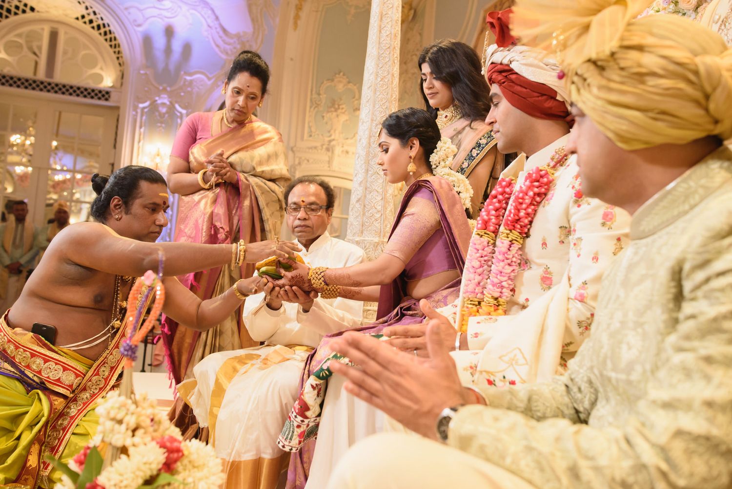 Hindu priest conducts sacred wedding rituals with family gathered around couple at ornate Indian wedding.