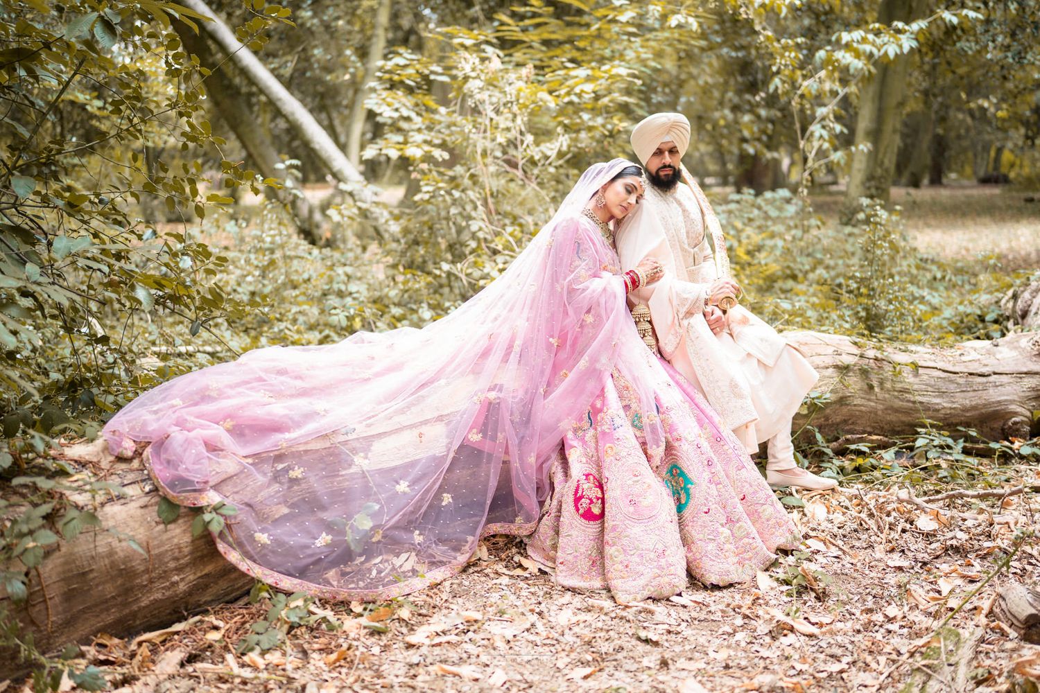 Bride and groom in pink Indian wedding attire pose by fallen log in forest setting.