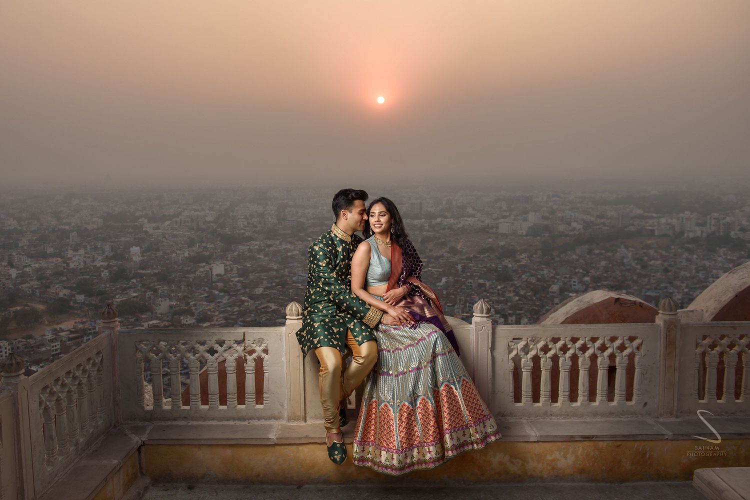 Couple in traditional Indian attire posing on a rooftop terrace at sunset with city views in Jaipur.