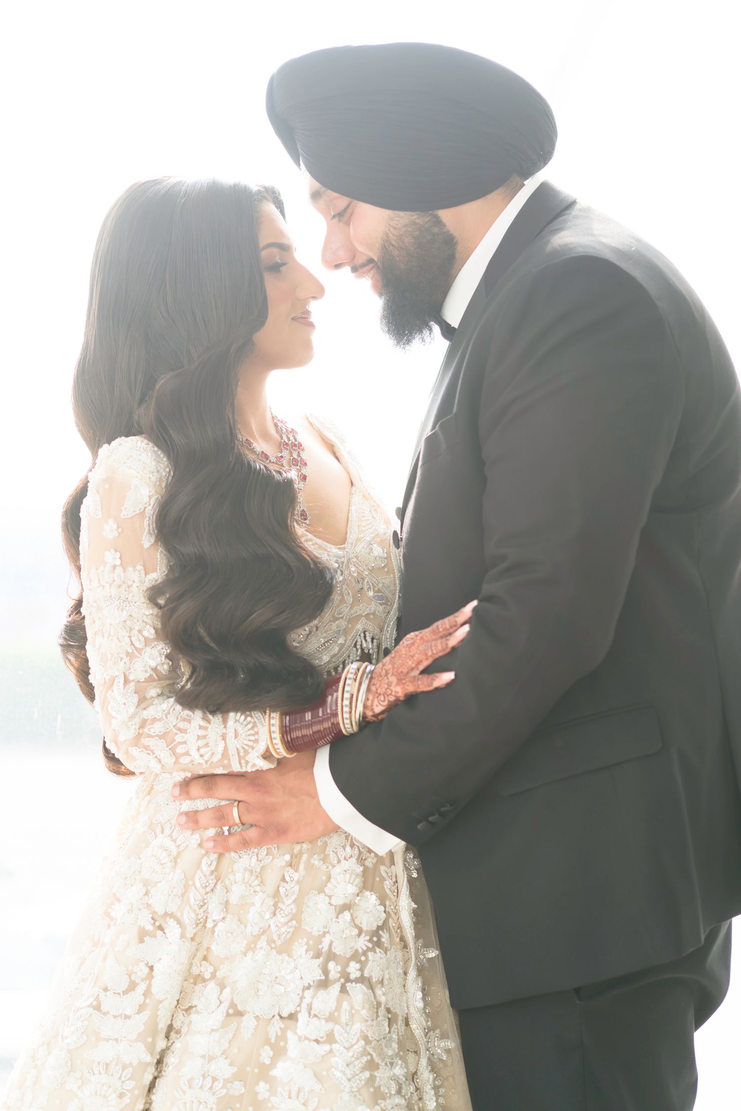 A Sikh groom in a black suit and turban tenderly embraces his bride in an ivory embroidered lehenga.