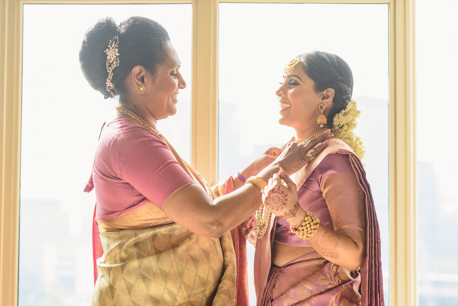 Indian mother helps bride with gold necklace jewelry while both wear traditional silk sarees.