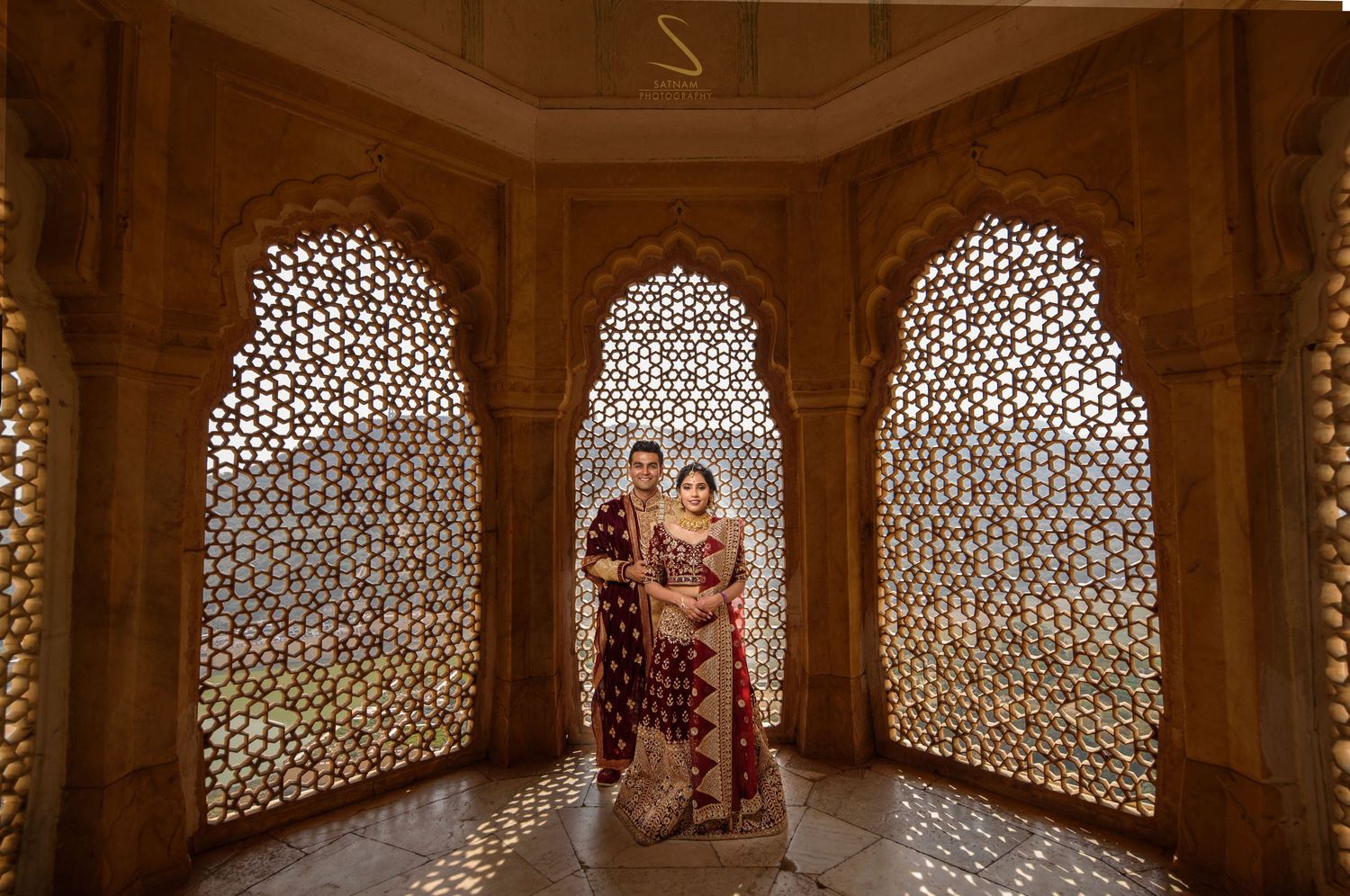 Indian wedding couple in maroon bridal wear posing inside ornate jali lattice pavilion with warm lighting.