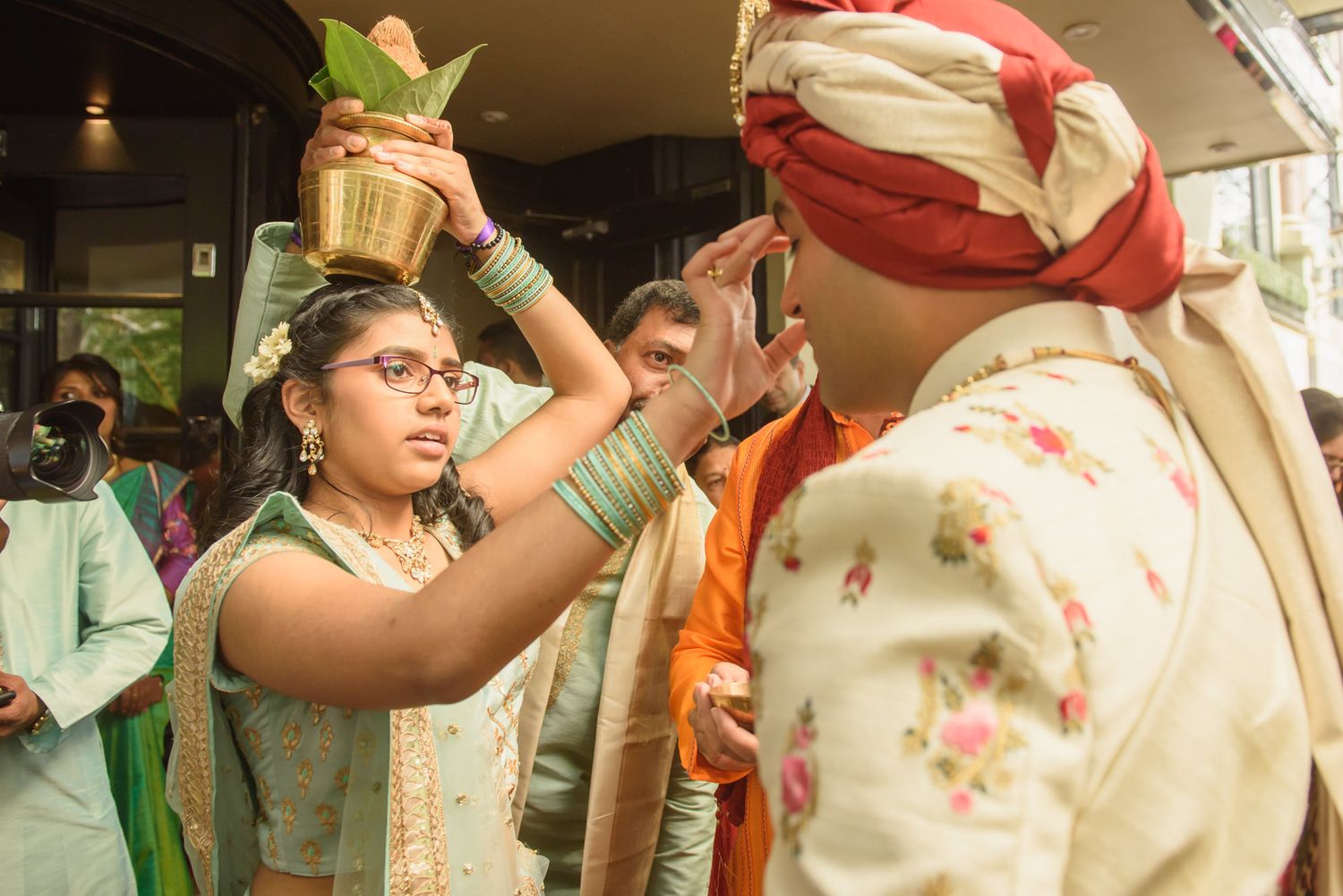 Woman performs traditional Indian wedding ritual pouring water from brass pot over groom's hands.