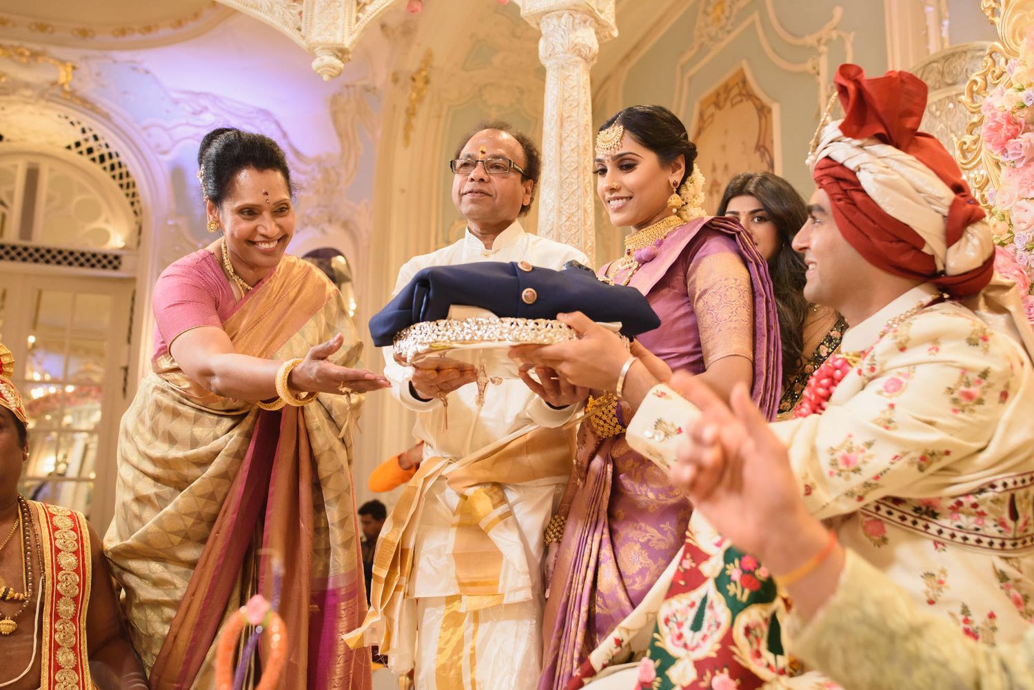 Family members present ceremonial tray to Indian bride and groom during traditional wedding celebration.