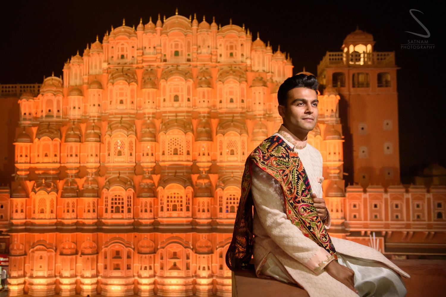 Groom in embroidered sherwani posing before illuminated Hawa Mahal at night in Jaipur.