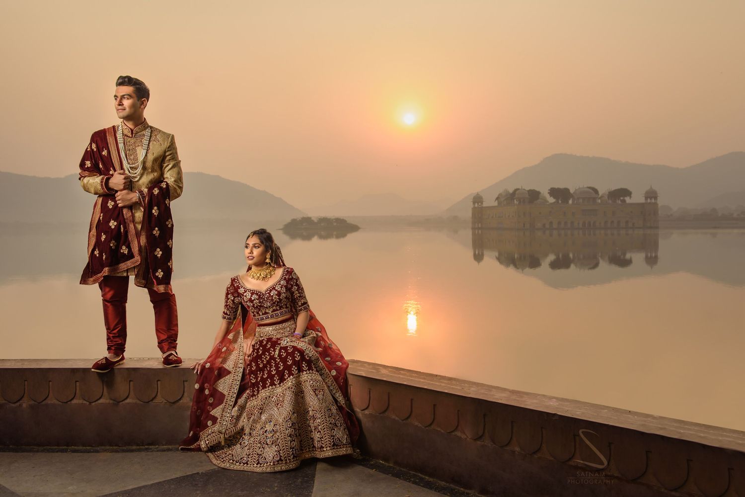 Indian bride and groom in maroon bridal attire posing by Jal Mahal palace lake at golden sunset.