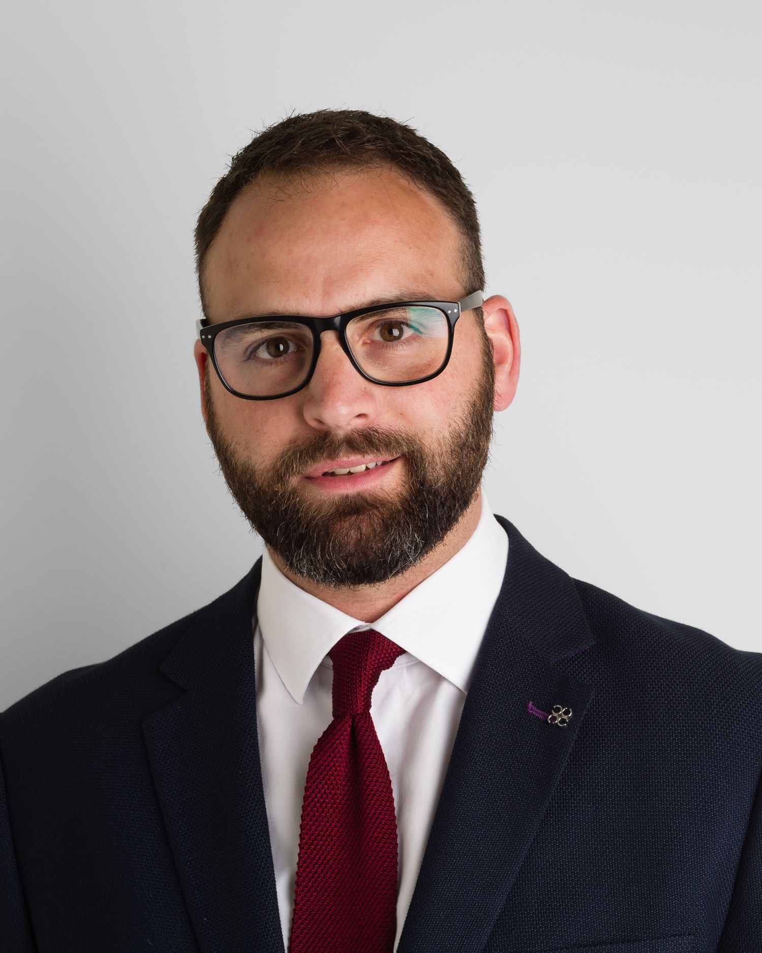Professional headshot of person in suit with glasses, beard and red tie.