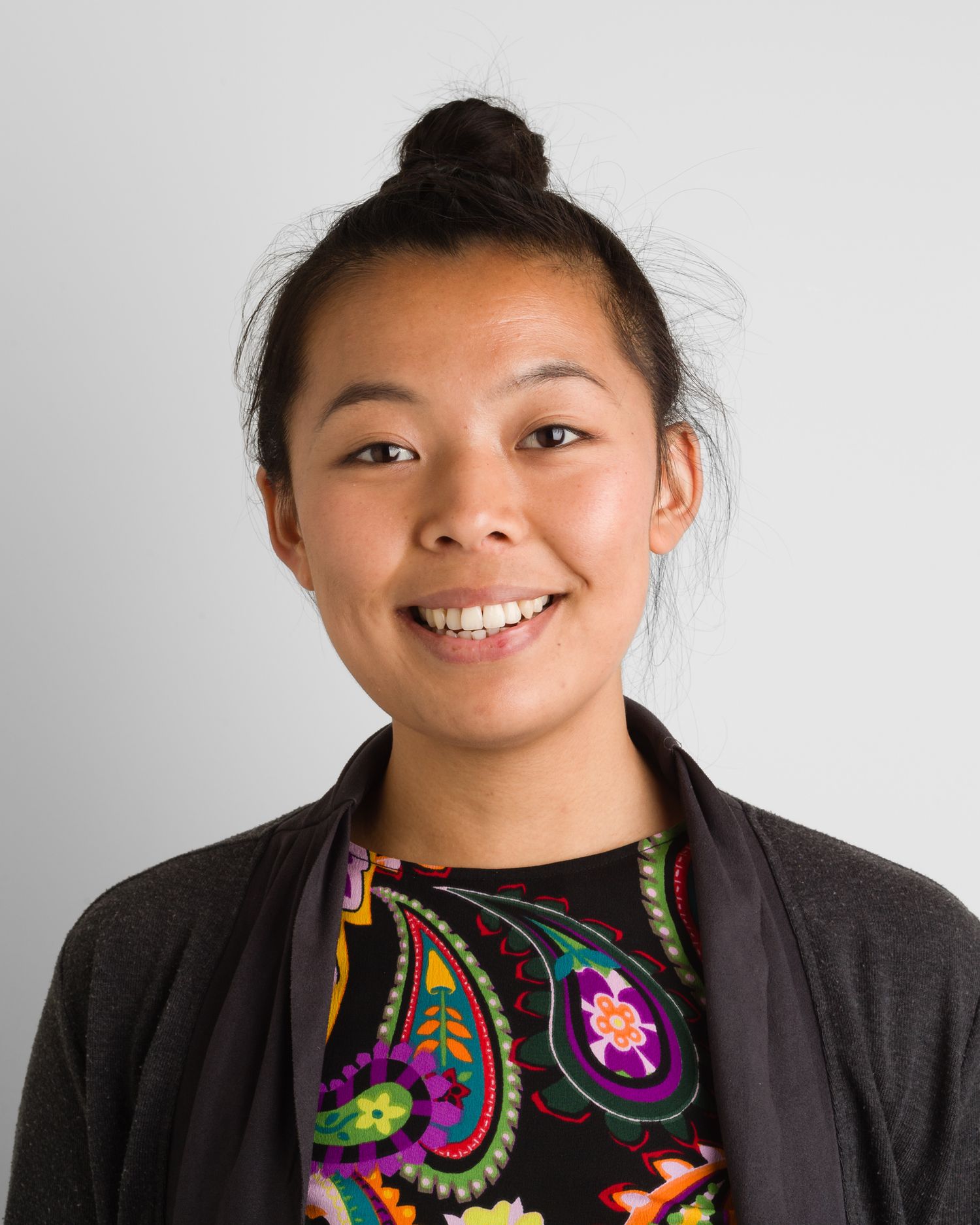 Business headshot Smiling person with hair in bun wearing patterned top and dark cardigan against light background.