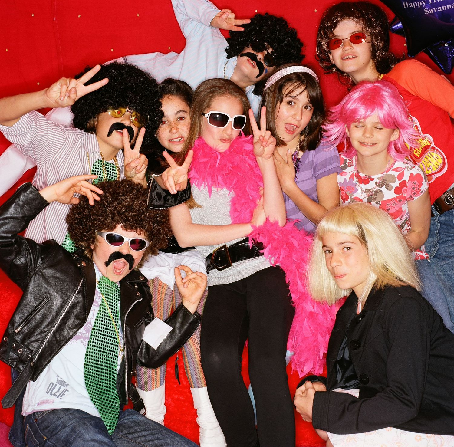 Group photo with party guests wearing sunglasses and fun accessories against red backdrop.