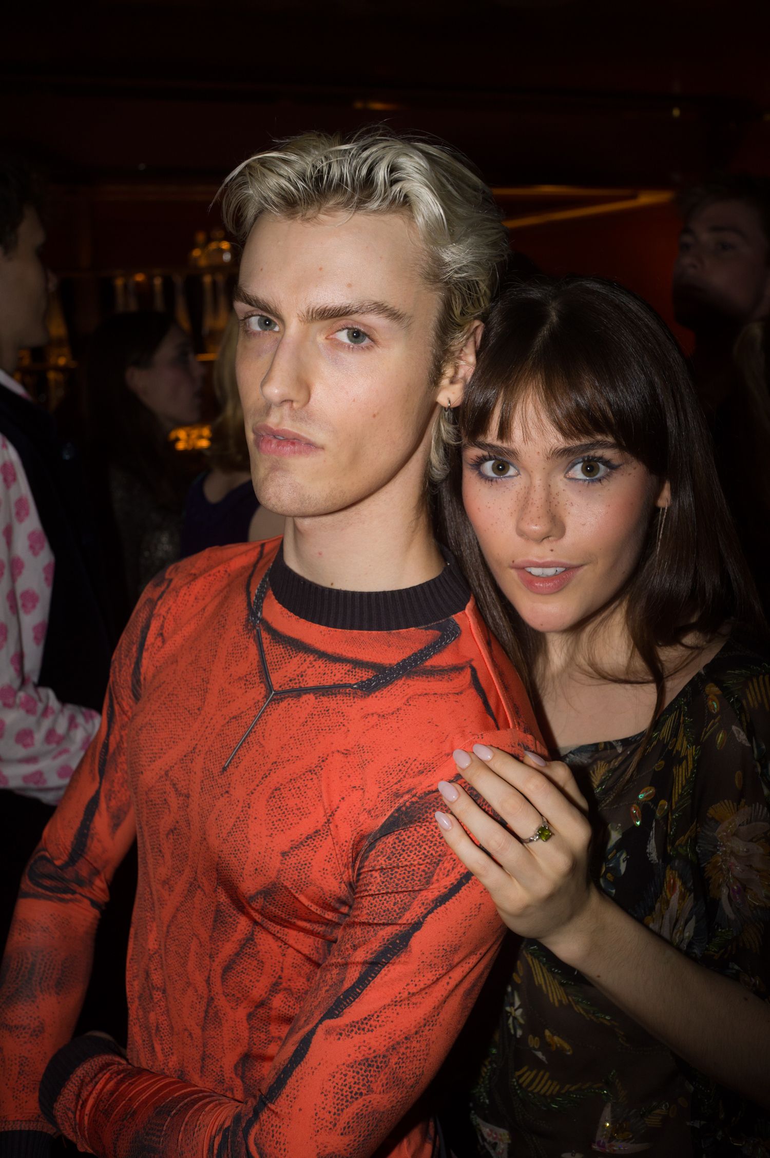 A couple in evening attire posing together at a party, with one wearing a red patterned shirt.