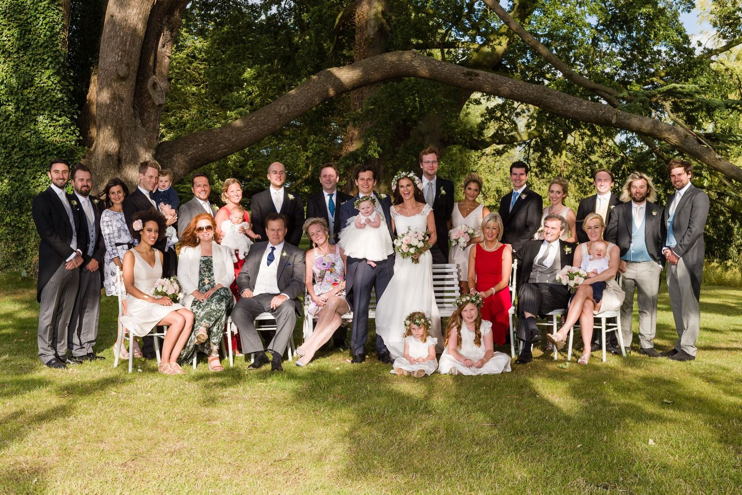 Large group photo of wedding guests gathered under trees in an outdoor garden setting.
