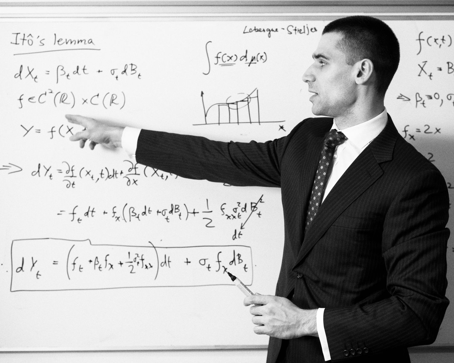 Mathematician in suit pointing at mathematical equations written on whiteboard.