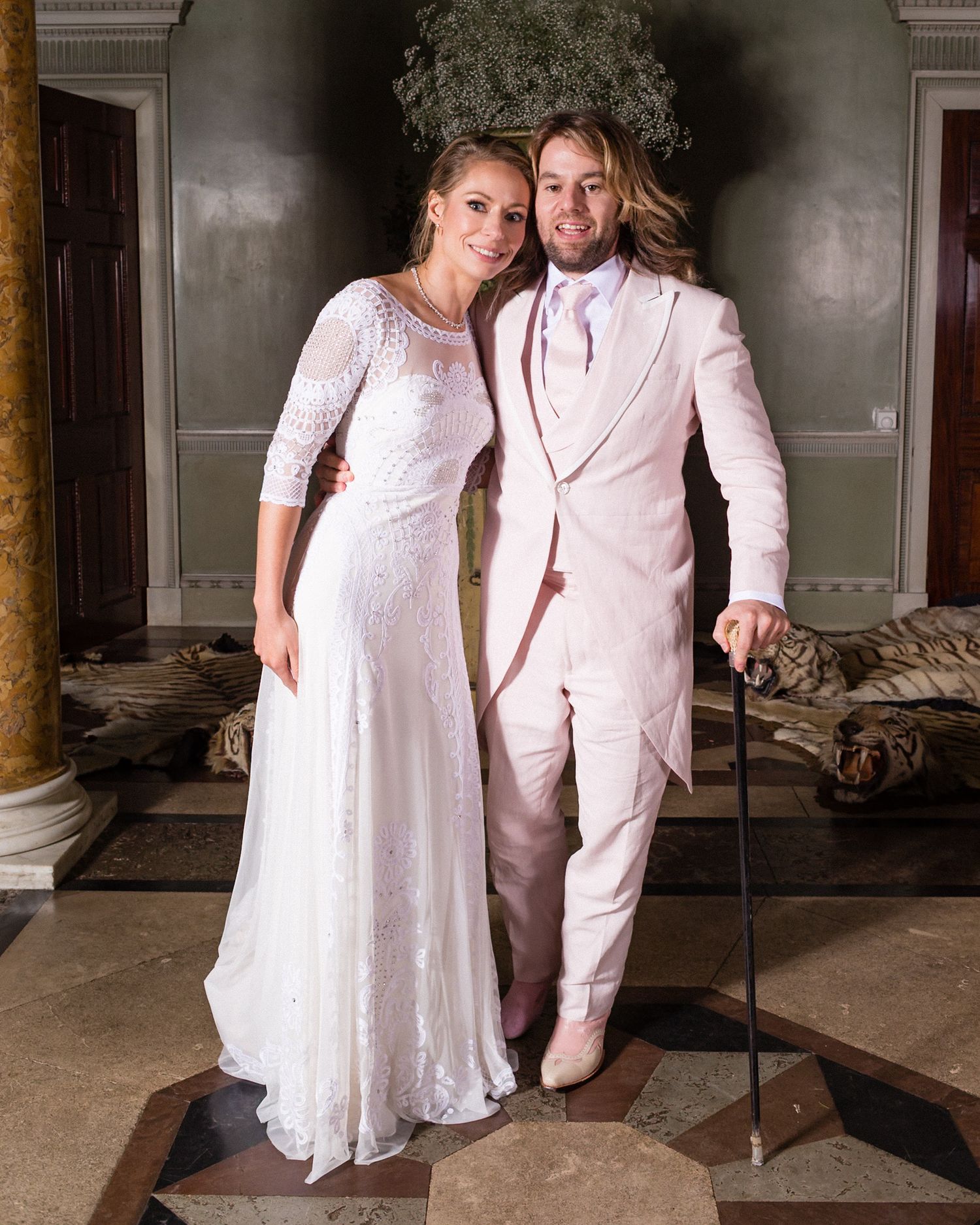 A couple poses in elegant white wedding attire against an ornate indoor backdrop.
