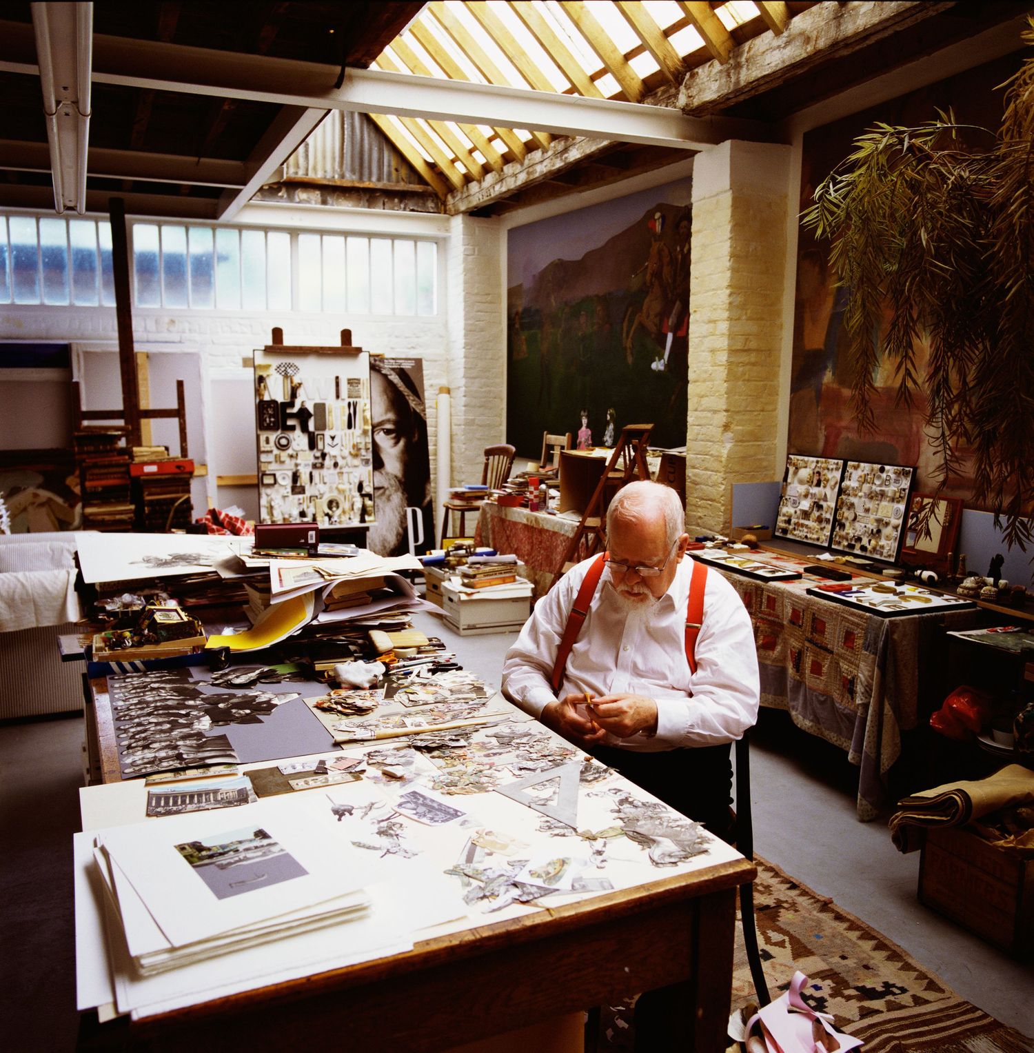Cluttered but tidy art studio workspace with tables covered in materials and supplies. Peter Blake seated at work.
