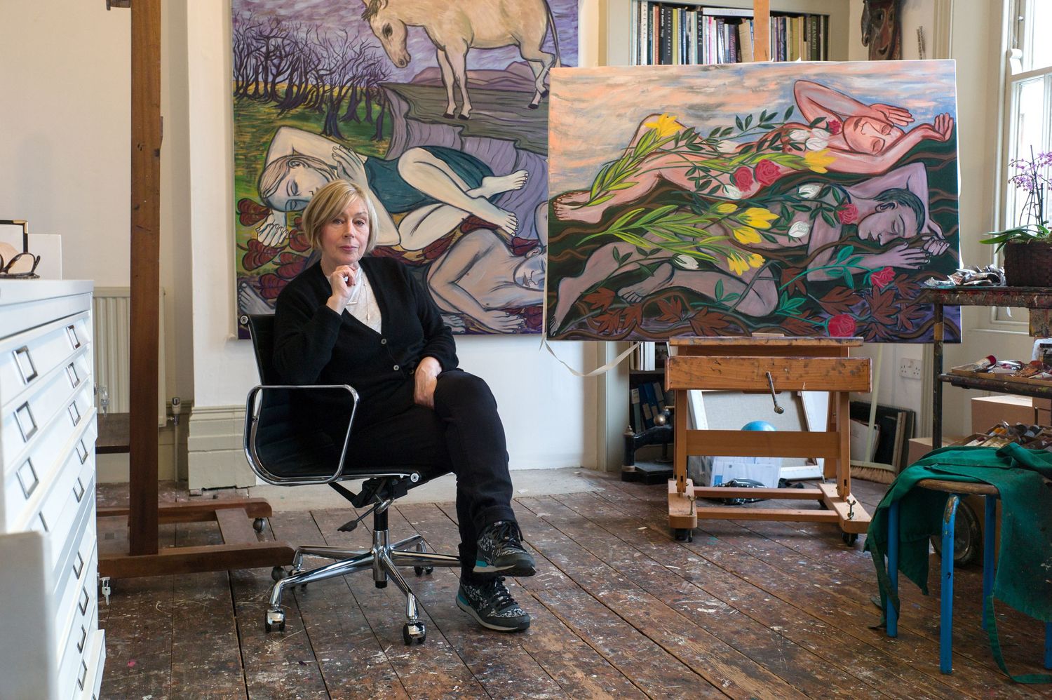 Artist working in a studio space with paintings and artwork displayed on walls and easels.