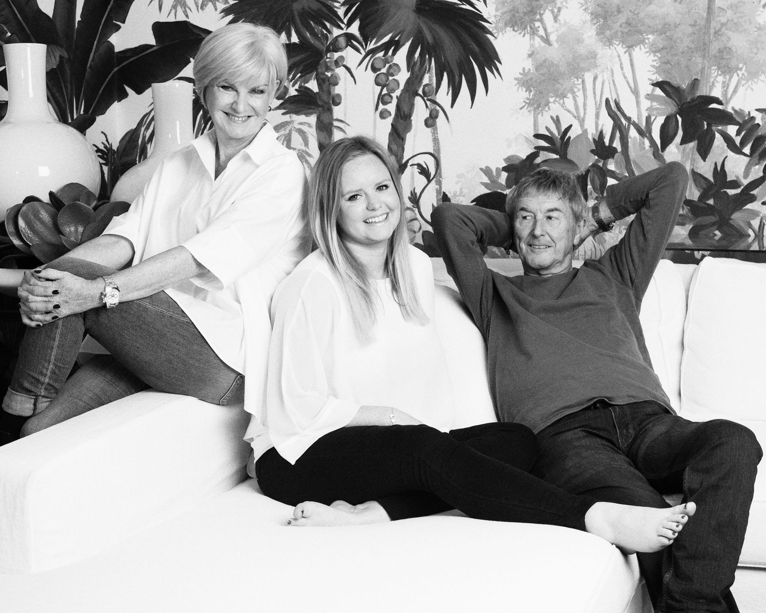Three people relaxing on white couch with tropical leaf wallpaper backdrop.