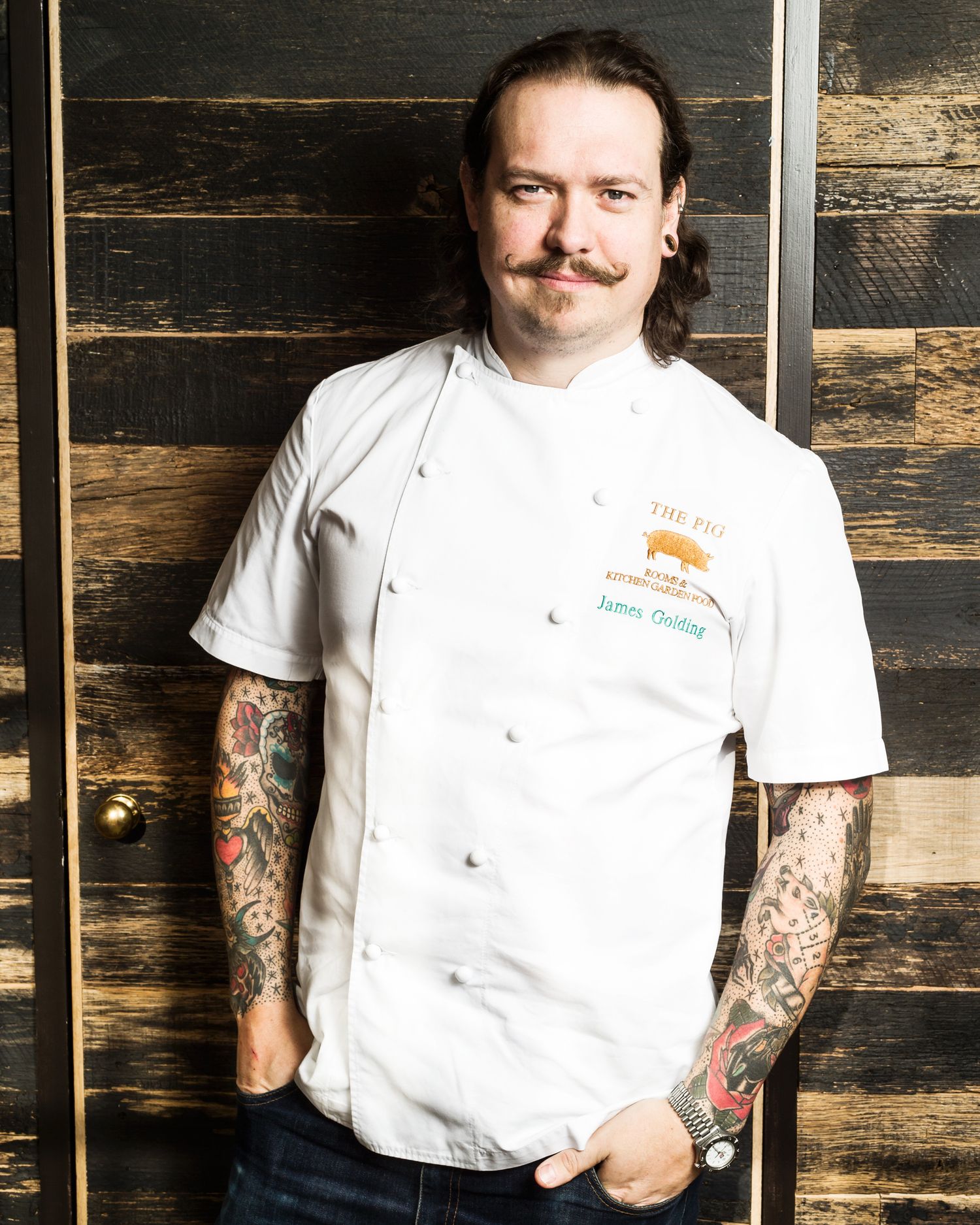 The Pig founder and Chef in white uniform with visible tattoos against rustic wooden wall background.