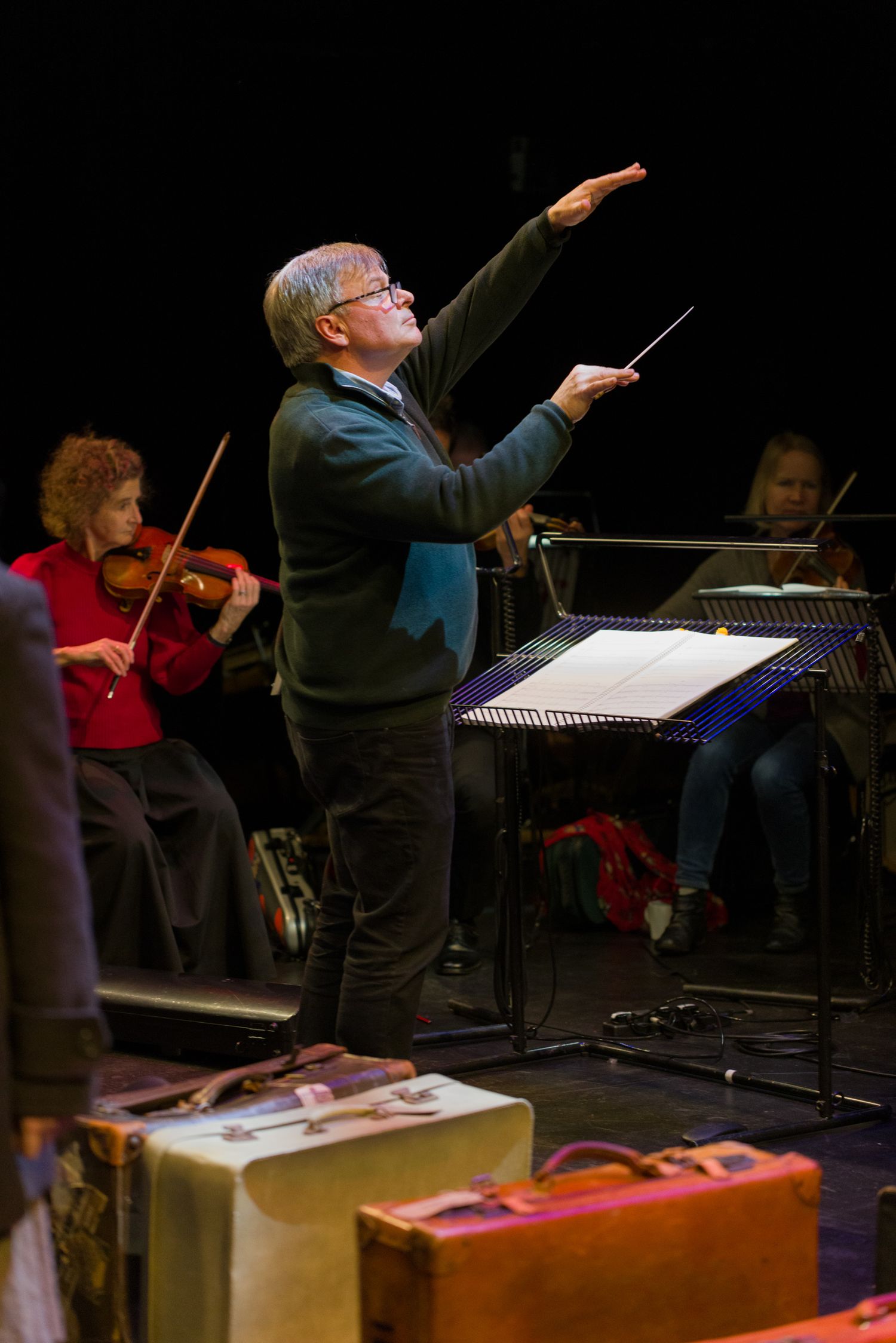 Orchestra conductor in dark attire directing musicians during performance on dark stage.
